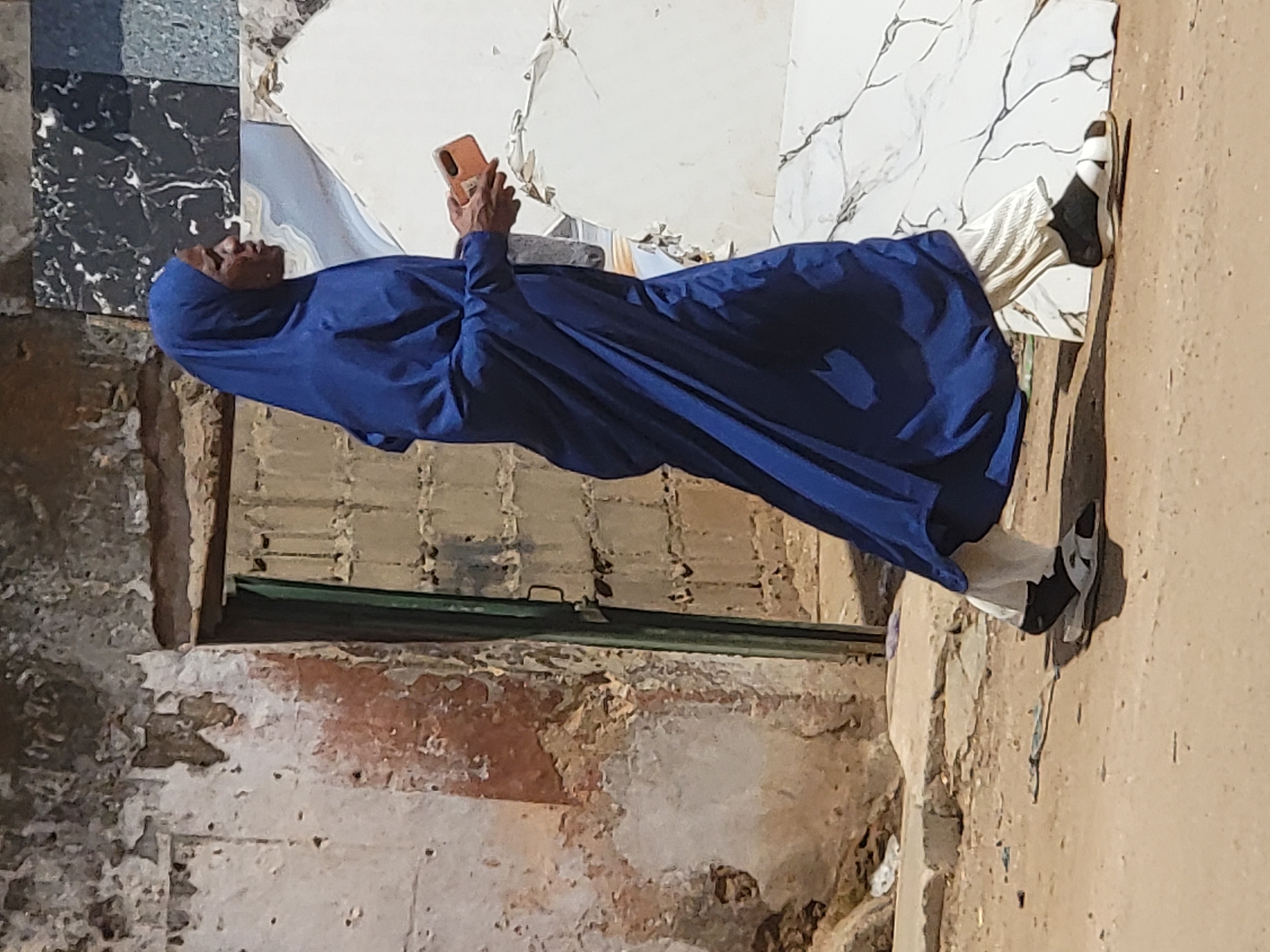 An elderly person wearing a blue hijab and a long blue garment is looking at a mobile phone, the phone's screen facing away. The person is also wearing white trousers and black-and-white sneakers. The person is standing or leaning against a wall which appears to be the exterior of a building. The building's exterior is weathered, with visible signs of aging and disrepair. Adjacent to the person is a wall with what looks like a combination of marble tiles and a door with a horizontal green bar. The setting is Maiburiji, Nigeria.