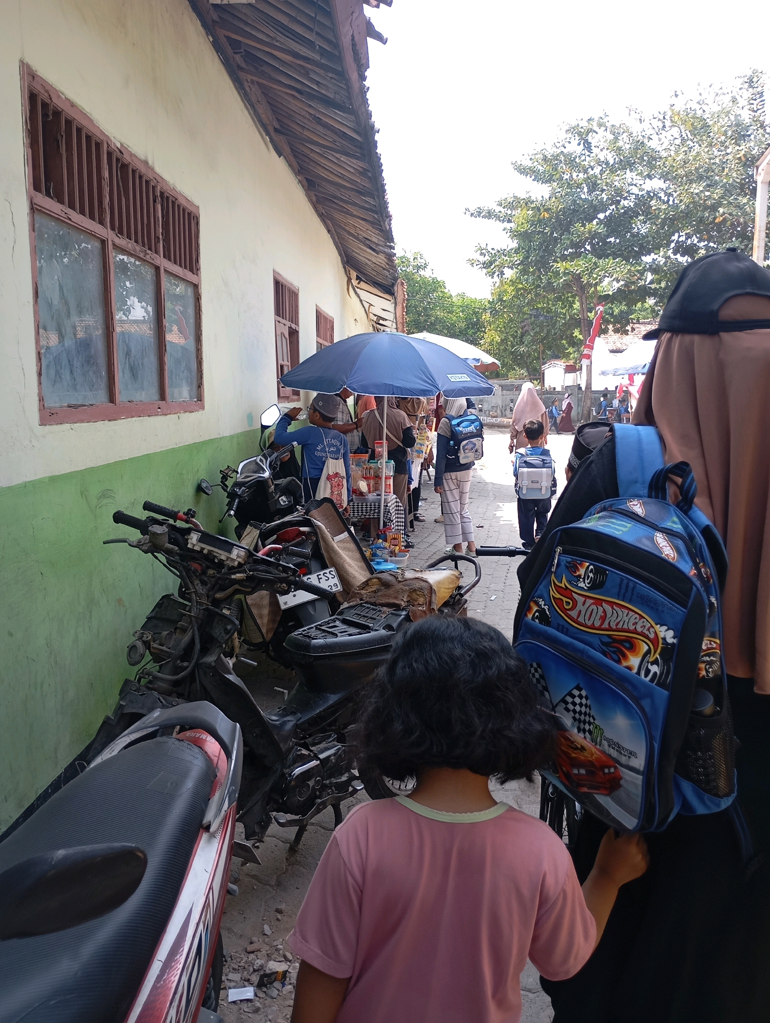 In a street scene in Babelan, Indonesia, several people are visible. A young child wearing a pink shirt stands in the foreground, facing away from the camera and reaching out towards a blue backpack with a Hot Wheels design. Next to the child, a dark-haired person, likely an adult, is also wearing dark clothing. In the background, more individuals are seen, some carrying backpacks, and several motorcycles are parked along a light-colored building with green trim. A blue umbrella is open, providing shade. The environment appears to be a marketplace or a bustling street with shops or stalls. The building's architecture includes wooden window frames with bars and a corrugated roof. The ground is dusty and strewn with small debris. The overall atmosphere suggests a daytime activity, possibly a school or a gathering.