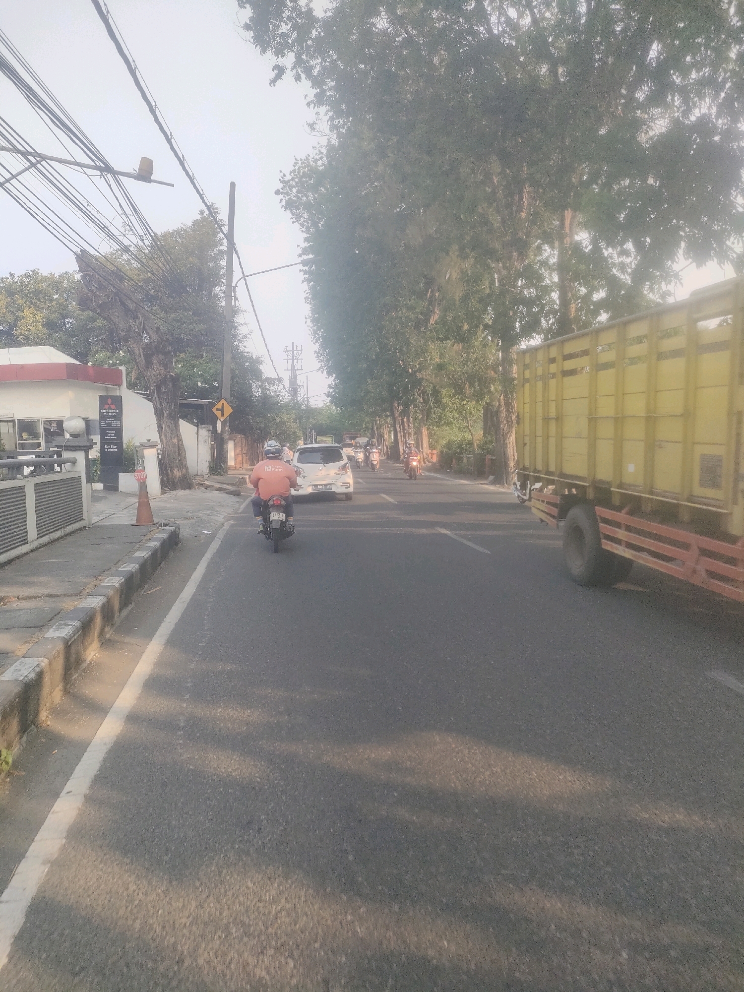 The image depicts a street scene in Surabaya, Indonesia, on a hazy day. A motorcycle with a rider wearing an orange shirt is in the foreground, traveling on the right side of the road. A white car is visible further ahead, also in the right lane. On the left side of the road, there is a building with signage that appears to be a car dealership, possibly Mitsubishi. A yellow truck is prominently featured in the right lane of the road, traveling in the same direction as the motorcycle. Trees line the sides of the road, and overhead power lines are visible. The overall impression is of a typical urban street with traffic and pedestrian activity.