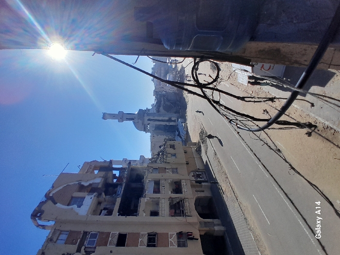 The image depicts a scene of significant damage and destruction in what appears to be an urban or semi-urban area, captured under bright, sunny conditions. The perspective is tilted, suggesting it was taken from a height or at an unusual angle, but if rotated clockwise to an upright position, it shows a street running roughly from the bottom-left to the top-right, flanked by buildings.
On the left side of the street, a multi-story building stands heavily damaged, with large sections of its facade torn away, exposing interior rooms and structural elements. Debris and rubble are visible, and several windows are broken or boarded up. Further down the street, towards the center-right, a mosque or similar religious building with a distinct minaret is visible, appearing largely intact, though parts of its structure might also show damage upon closer inspection. Other buildings, some partially obscured or appearing damaged, line the street in the background.
The sky is a clear, bright blue, with the sun prominently visible in the upper-left corner (when upright), emitting strong rays and lens flare. Numerous black electrical wires or cables are strung across the upper portion of the frame, partially obscuring the view of the sky and some buildings. The road itself is asphalt, with a white dividing line, and appears largely empty of traffic or people. The ground adjacent to the road is sandy or dusty.
No specific activity is clearly depicted, but the extensive damage to the buildings strongly suggests the aftermath of conflict or a major destructive event. The time of day appears to be midday or early afternoon due to the high, bright sun. In the bottom-right corner, the text "Galaxy A14" is visible, indicating the device used to capture the image. The scene conveys a sense of devastation and resilience under a clear sky.