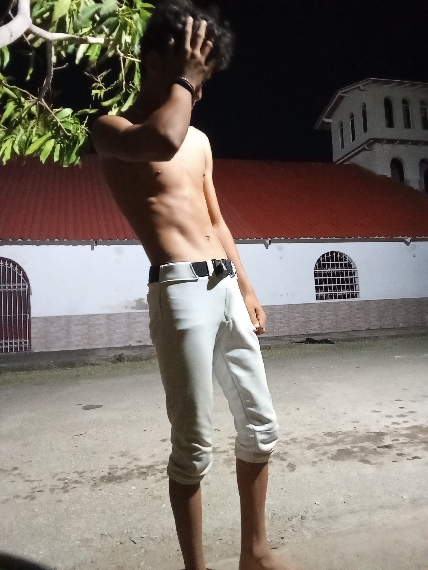 The image shows a shirtless young man standing barefoot on a paved surface at night. He is wearing light-colored pants with a black belt. His right hand is on his head, obscuring his face.
In the background, there is a white building with a red roof and arched windows, possibly a church. A tree with foliage is visible on the left side of the frame. The image appears to be taken outside at night, with limited lighting.
The location of the image is consistent with the city of Juan Griego, Venezuela, as churches with similar architectural features are located in this city.