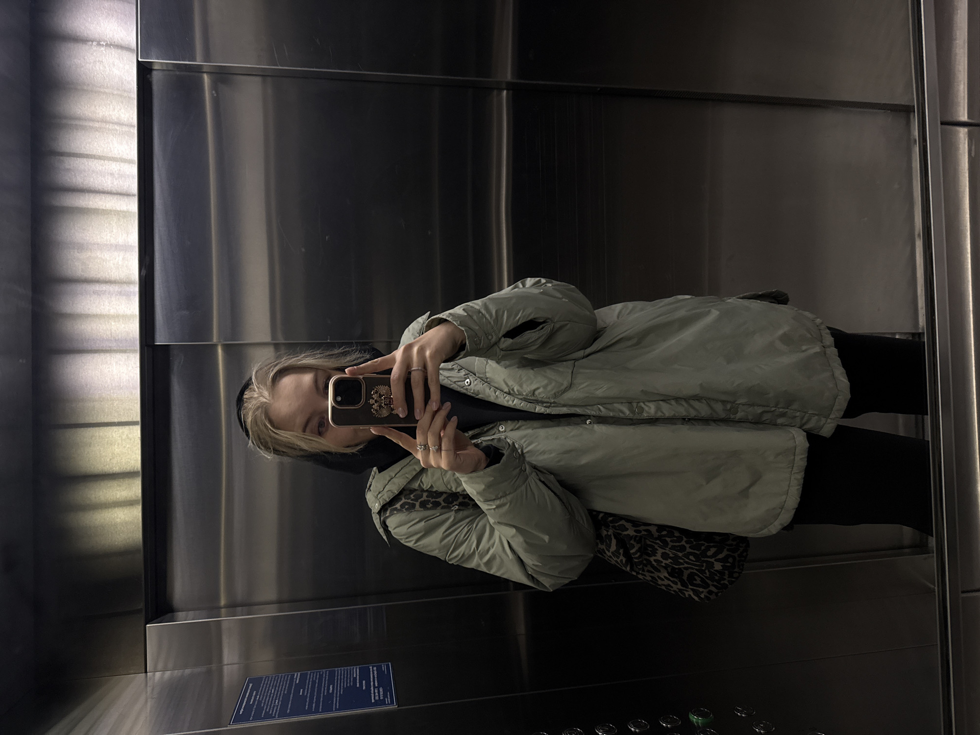 A person is captured in a mirror selfie inside an elevator. The image is oriented sideways, with the person appearing to lie horizontally across the frame.
The central subject is an individual, likely female, with blonde hair that frames their face and light-colored eyes. They are wearing a light green or mint-colored puffer jacket over a dark-colored, possibly black, hoodie or top. Black trousers or leggings are visible below the jacket. Several rings adorn their fingers. The person is holding a smartphone with a distinctive case featuring a golden, intricately detailed emblem, which strongly resembles the Russian coat of arms (a double-headed eagle) on a dark background. A leopard print bag is slung across their body, visible beneath the jacket on their left side.
The setting is the interior of an elevator, characterized by highly reflective, possibly mirrored, walls that create a distorted reflection. The walls also feature brushed metal panels, suggesting a modern aesthetic. At the bottom of the reflection, part of the elevator control panel with multiple circular buttons is visible. On the lower right side of the mirrored wall, an information placard with text in Cyrillic script is affixed, though the text itself is unreadable due to distance and focus.
The activity taking place is the person taking a mirror selfie, looking at the screen of their phone. The overall lighting is dim, suggesting an indoor environment, typical of an elevator. The provided context indicates the location is Krasnoyarsk, Russia.