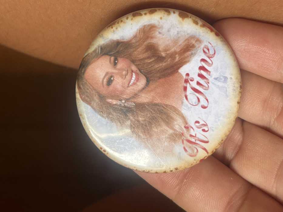 A human hand with light brown skin is shown in a close-up, holding a circular pin or button. The button, approximately 4-5 cm in diameter, features a printed image of Mariah Carey, a light-skinned female with long, wavy blonde hair, smiling directly. She is adorned with prominent, dangling earrings. Below her image, the text "It's Time" is written in a decorative red script font. The button's surface is worn, exhibiting multiple fine scratches and scuffs, and its outer edges display irregular discoloration and browning. The fingers of the hand are gripping the button's rim, presenting its face. The background behind the hand is dark, out of focus, and indistinct, providing no discernible environmental features. The specific location within Buenos Aires, Argentina, cannot be visually identified from the scene depicted.