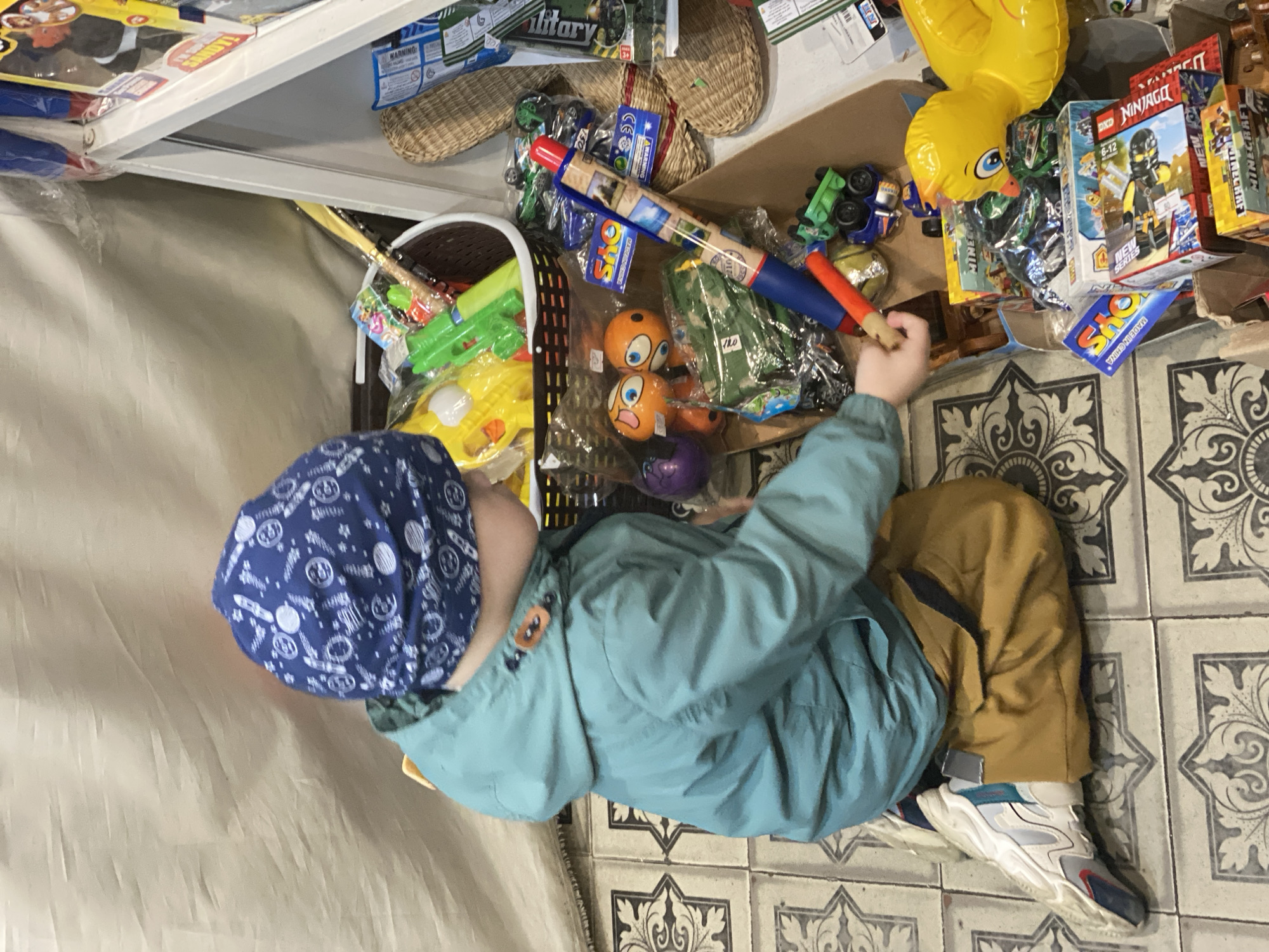A young child is observed kneeling on an ornate tiled floor, facing away from the viewer, in a retail environment in Sochi, Russia. The child wears a blue beanie with white celestial patterns, a teal hooded jacket with visible brown buttons, mustard-colored pants, and white, grey, and blue athletic sneakers. Their right hand is extended towards a blue and red toy rocket or missile, situated among a densely packed collection of toys. To the child's immediate left, a brown plastic basket contains various packaged toys, including yellow inflatable objects and green water guns. To the right, a white shelving unit is partially visible, and the floor area is covered with a variety of packaged toys, some identified as "Minecraft" and "NINJAGO," small green and blue toy vehicles, and woven wicker items. A light tan fabric curtain serves as the backdrop to the scene, suggesting a market stall or informal shop setting.