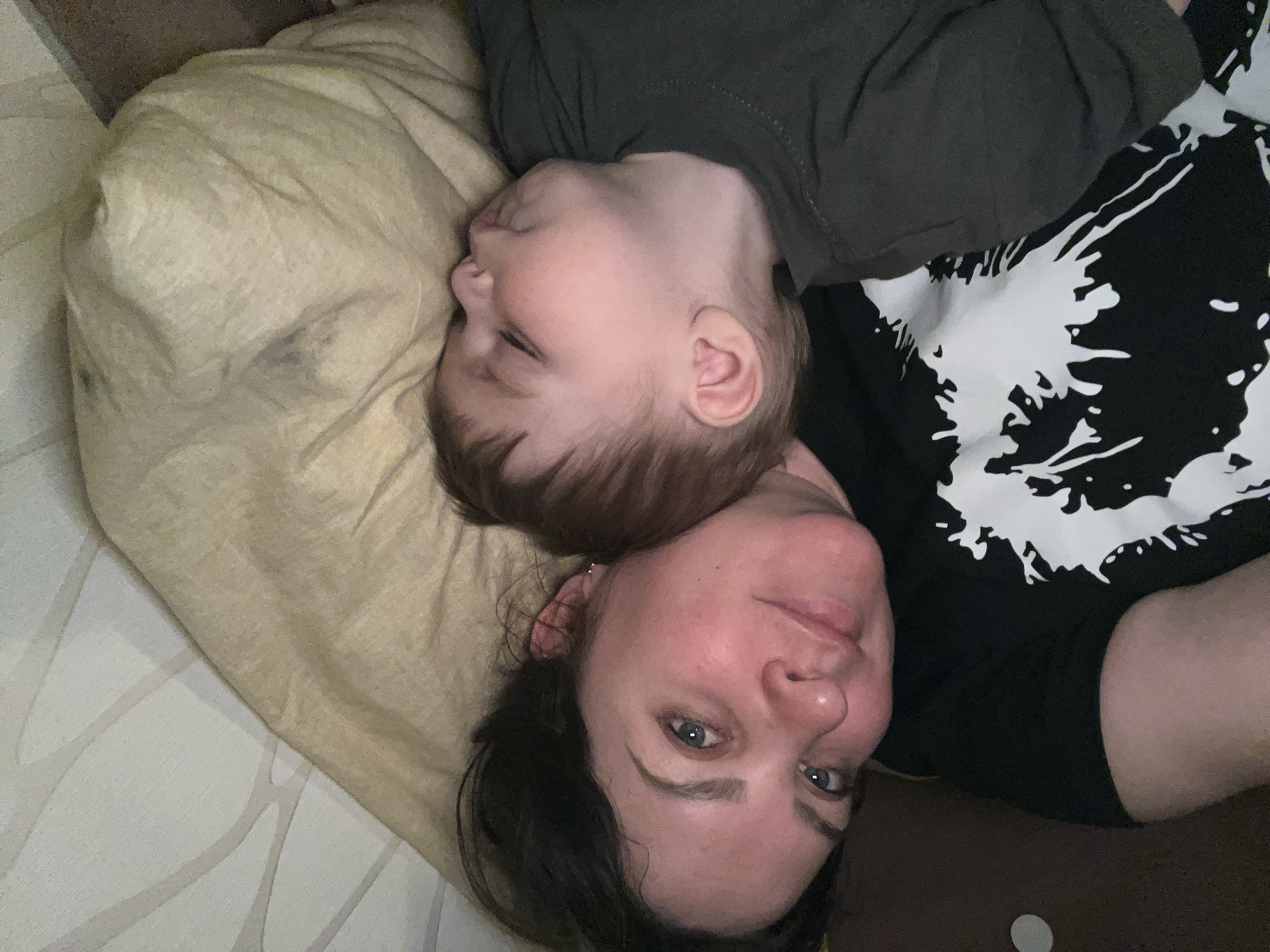 A woman and a child are lying on a bed. The woman is facing the camera, and the child is lying next to her, with the child's head on the woman's chest. The woman has dark hair and blue eyes, and she is wearing a black t-shirt with a white design. The child has brown hair and is wearing a dark gray shirt. The bed has a beige-colored pillow and a white and gray patterned sheet. The photo was taken indoors, likely in a home. There is no indication of the city of Perm, Russia, in the image.