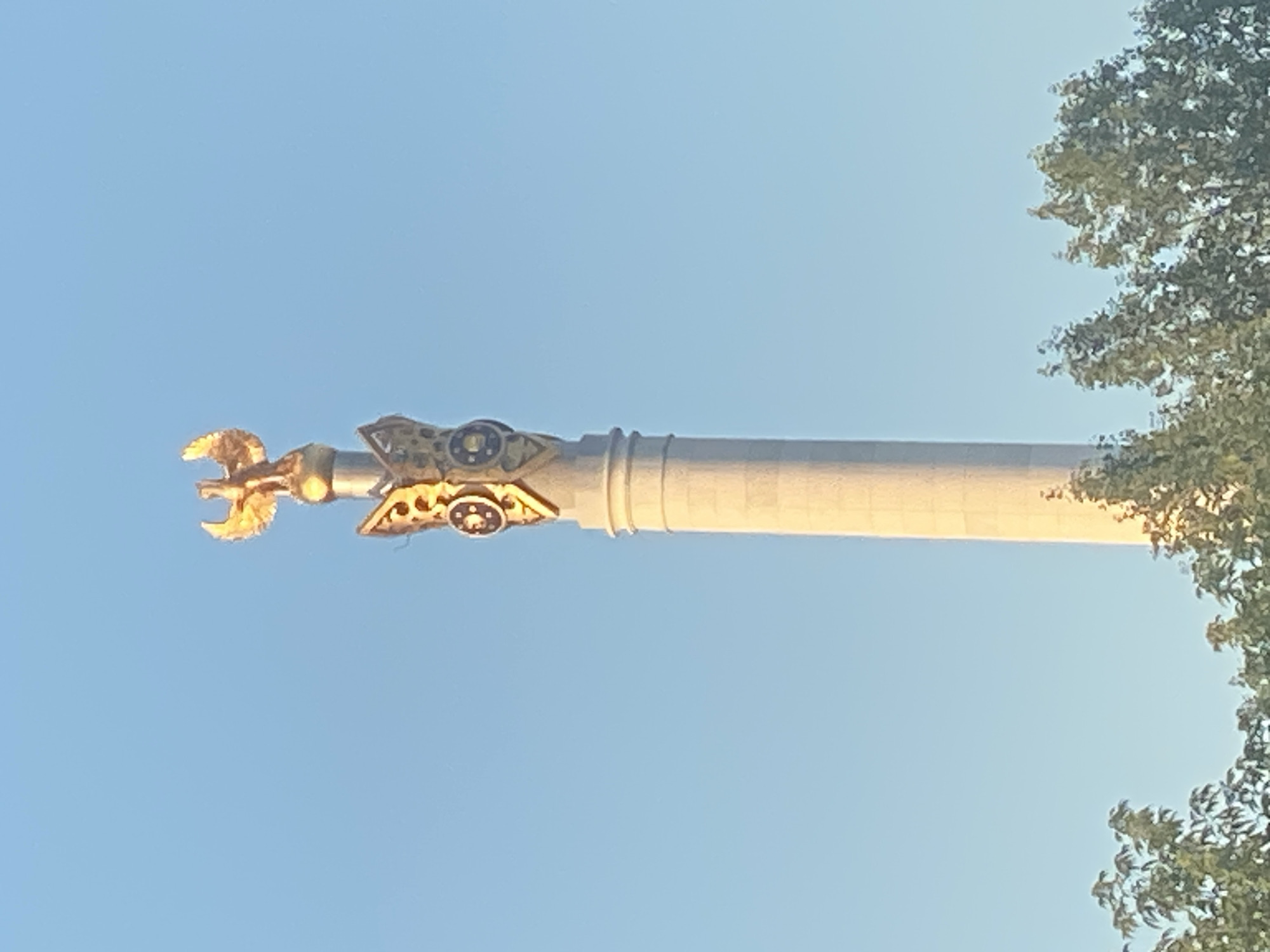 The image shows the top portion of the Bayterek Tower in Astana, Kazakhstan. The tower is a monument and observation tower, and its top is adorned with a golden eagle sculpture. The sky is clear and blue, with some trees visible at the bottom of the frame. The golden eagle appears to be a prominent feature, with its wings spread. The tower itself is tall and cylindrical, with decorative elements near its apex. The lighting suggests it is daytime, possibly late afternoon or early morning, given the warm glow on the golden sculpture.