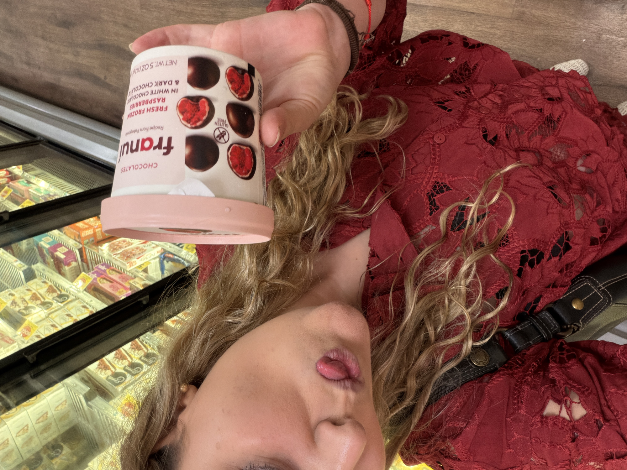 A woman in a red lace top is holding a container of Franui chocolates. The container has the brand name "Franui" printed on it in large pink letters, followed by "CHOCOLATES". Below the brand name, it says "Recipe from Patagonia" and "FRESH FROZEN RASPBERRIES IN WHITE CHOCOLATE & DARK CHOCOLATE". The net weight is listed as "NET WT. 5 OZ (142g)". The container's design features images of the chocolate-covered raspberries. The woman is in front of a refrigerated case filled with frozen desserts. This scene likely takes place inside a grocery store. While specific geographical information is not readily available from the image, the general geographical setting is Key Biscayne, United States of America. The specific location can not be determined from the given information.