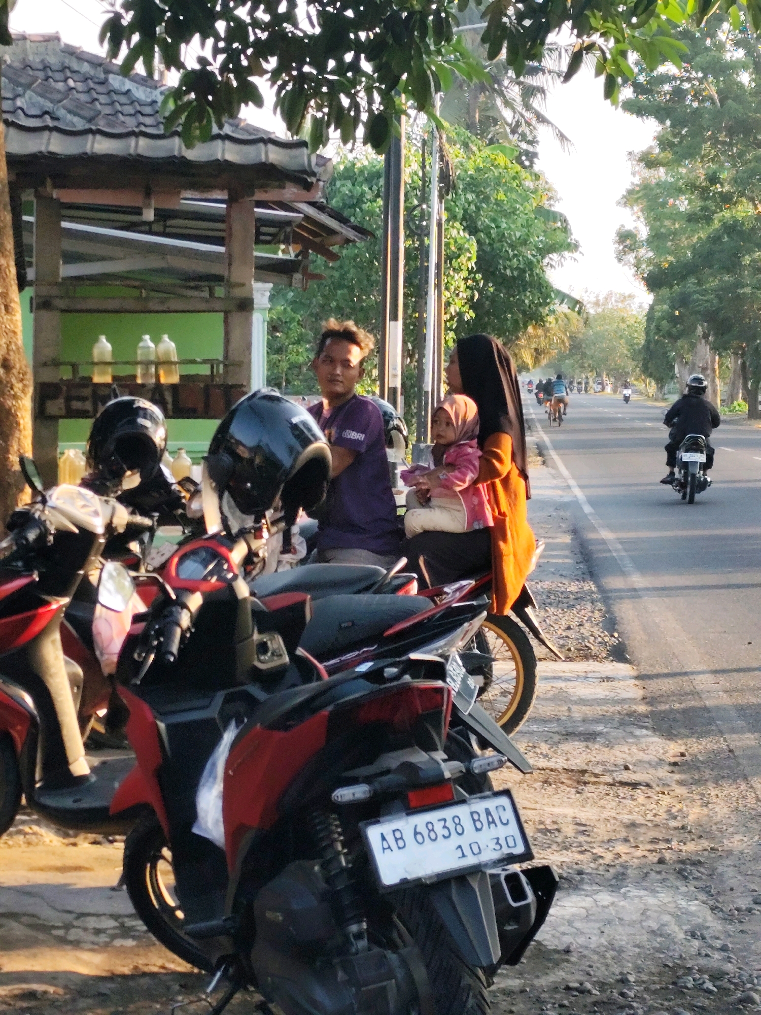 In the city of Bambang lipuro, Indonesia, a man stands beside a row of motorcycles. He is wearing a purple t-shirt and has short, messy hair. To his right, a woman in a black hijab and an orange dress holds a baby dressed in a pink jacket. The woman is seated on a motorcycle, and the baby is on her lap. Two black helmets are placed on the handlebars of motorcycles in the foreground. Behind the people, a structure with a tiled roof is visible, and three plastic bottles filled with yellow liquid are displayed on shelves. The background shows a street with several motorcycles and people, suggesting a busy area. Trees line the street, and the sunlight casts shadows on the ground, indicating it is either morning or late afternoon. The motorcycles are parked along a paved road, with a gravel shoulder. The license plate of one of the motorcycles in the foreground reads "AB 6838 BAC 10.30".