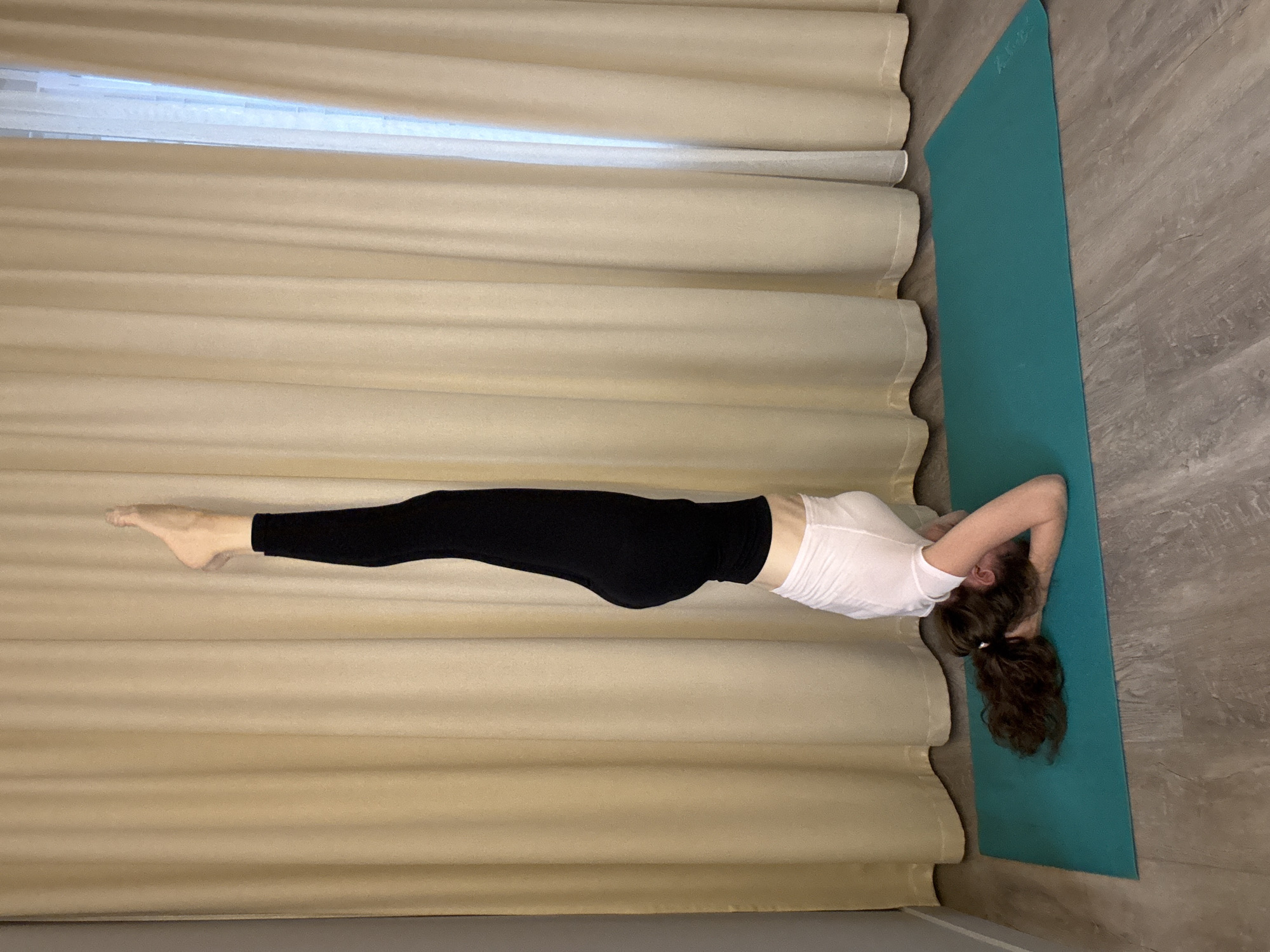 An individual is performing a headstand yoga pose on a turquoise yoga mat. The person is wearing a white t-shirt and black leggings, with their legs extended straight upwards. Their head is supported by their interlaced hands on the mat, and their hair is spread out around their head. The background consists of beige curtains and a white sheer curtain on the right, suggesting an indoor setting, possibly a home. The floor is made of wood-look laminate. The image does not provide any specific visual cues to determine its location within the city of Dolgoprudny, Russia.