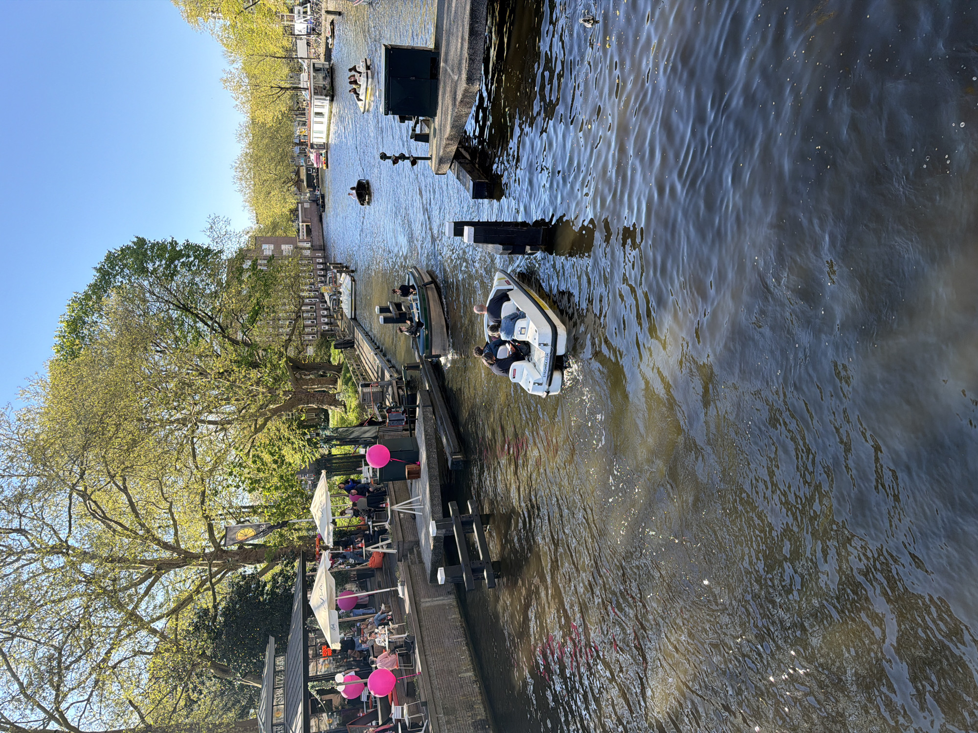 The image displays a lively canal scene in Amsterdam on a bright, sunny day, likely in spring or early summer, given the vibrant green foliage on the trees. The setting includes a wide canal bordered by lush trees and buildings, with an outdoor bar or restaurant area on one side.
Several subjects are visible throughout the image. People are actively engaged in leisure activities; some are seen navigating the canal in boats, while others relax at tables in the outdoor dining area. A prominent white motorboat with individuals onboard moves across the water in the middle ground, displaying "STRØMRA.COM" on its stern. Further along the canal, other boats, including a darker vessel carrying passengers, add to the maritime activity. Buildings with typical Dutch architecture line the canal banks, and houseboats are moored in various spots.
The outdoor bar area features white patio umbrellas providing shade over tables and chairs, which are occupied by patrons. Bright pink spherical balloons adorn the space, enhancing its festive atmosphere. A black flag with a yellow lemon slice and the word "SPRITZ" is visible, along with a sign on the building structure that reads "APERO SPRITZ BAR," clearly indicating the establishment's type.
The water of the canal is dark but reflects the clear blue sky and sunlight, creating shimmering patterns. The weather is fair, with bright sunshine suggesting a pleasant day for outdoor activities. The scene conveys a relaxed and enjoyable atmosphere typical of a leisurely day in a city known for its canals.