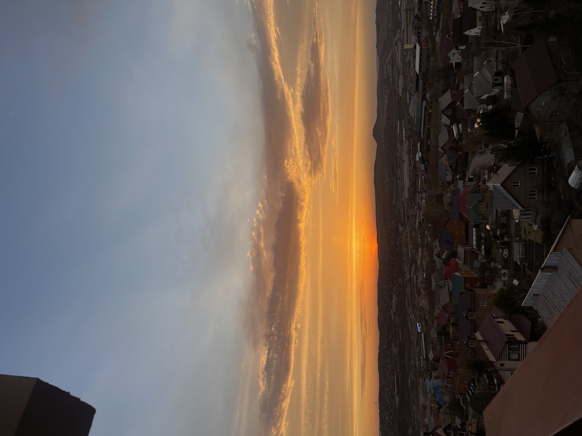 The image captures a sunset over Krasnoyarsk, Russia. In the foreground is a neighborhood of houses with various colored roofs. The skyline is punctuated by the outline of mountains in the distance. The sky is filled with a mix of blue and orange hues as the sun sets, with textured clouds stretched horizontally across the frame. The image is shot from an elevated perspective.