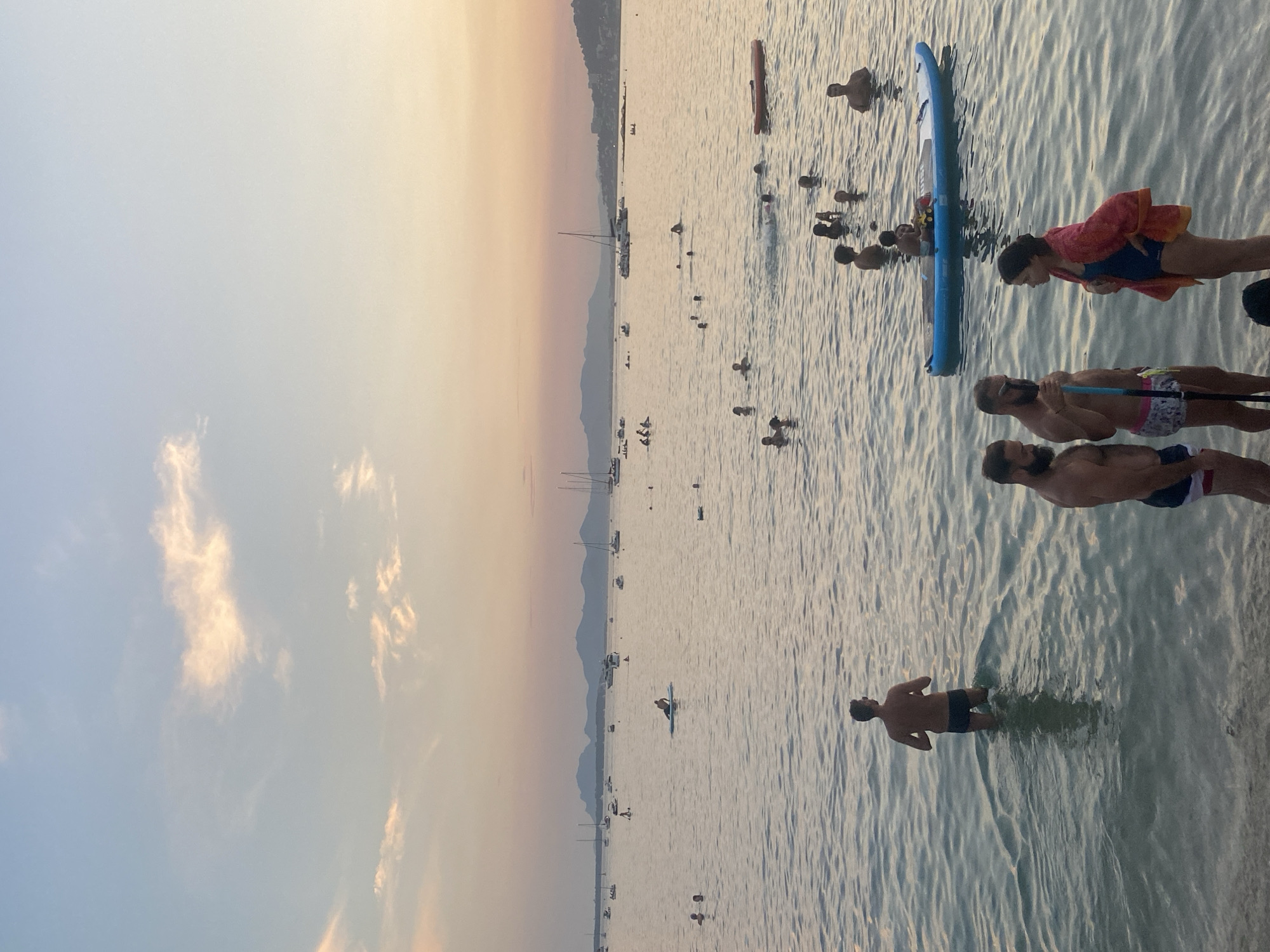 The image depicts a serene coastal scene at dusk, likely in Antibes, France. The sky is a gradient of soft blues and oranges, with a few wispy clouds. The sea is calm, reflecting the sky's colors, and dotted with numerous boats, including sailboats and smaller vessels, anchored in the distance. In the foreground, several people are in the water. Some are swimming, while others are standing or wading near the shore. One person is on a paddleboard, and another individual is seated on it. A group of three people are conversing near the shore, two men and a woman wearing a towel draped over her shoulders. In the mid-ground, a man stands in the water, facing away from the camera, and another man is visible in the shallow water at the bottom edge of the frame. The overall atmosphere is one of relaxation and leisure as the day comes to a close.