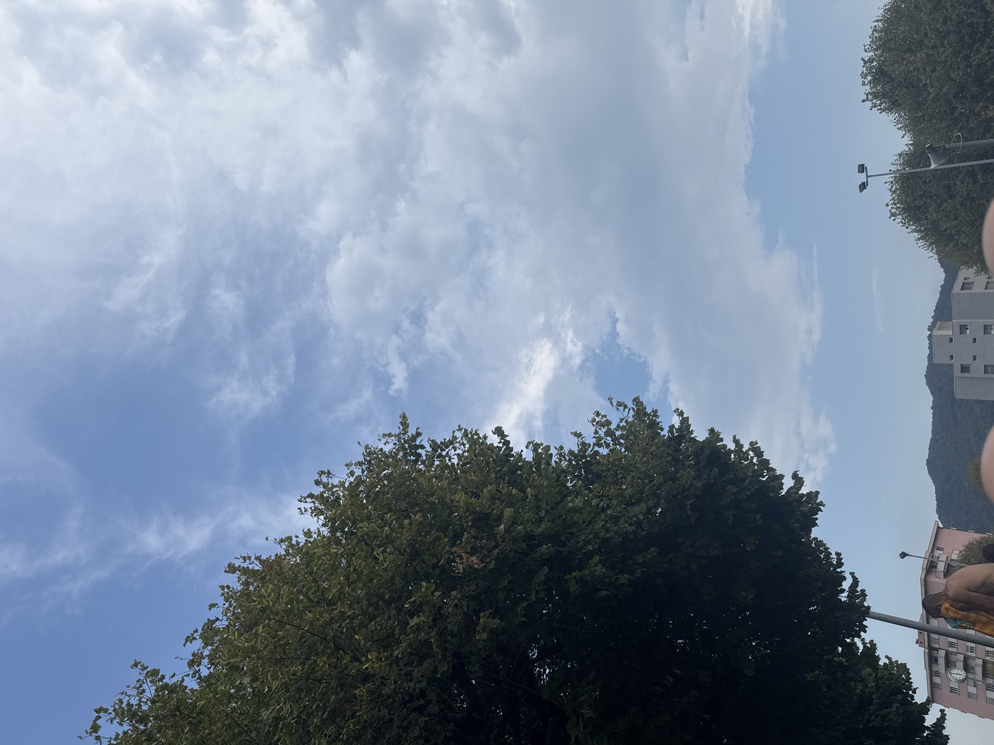 An outdoor, low-angle, full shot depicts the sky above buildings and trees in Chiasso, Switzerland. The sky is mostly blue with scattered white clouds. On the left side of the frame, a large tree with dark green leaves is prominent. In the lower left quadrant, a shirtless man is visible from the waist up, drying himself with a towel. Behind him, a pink multi-story building with a clock on its facade is partially obscured by the tree. To the right of the man and building, other buildings and lush green trees are visible, with hills in the background. A street lamp with two lights stands in the middle ground. The overall scene suggests a casual moment outdoors, possibly near a residential area.