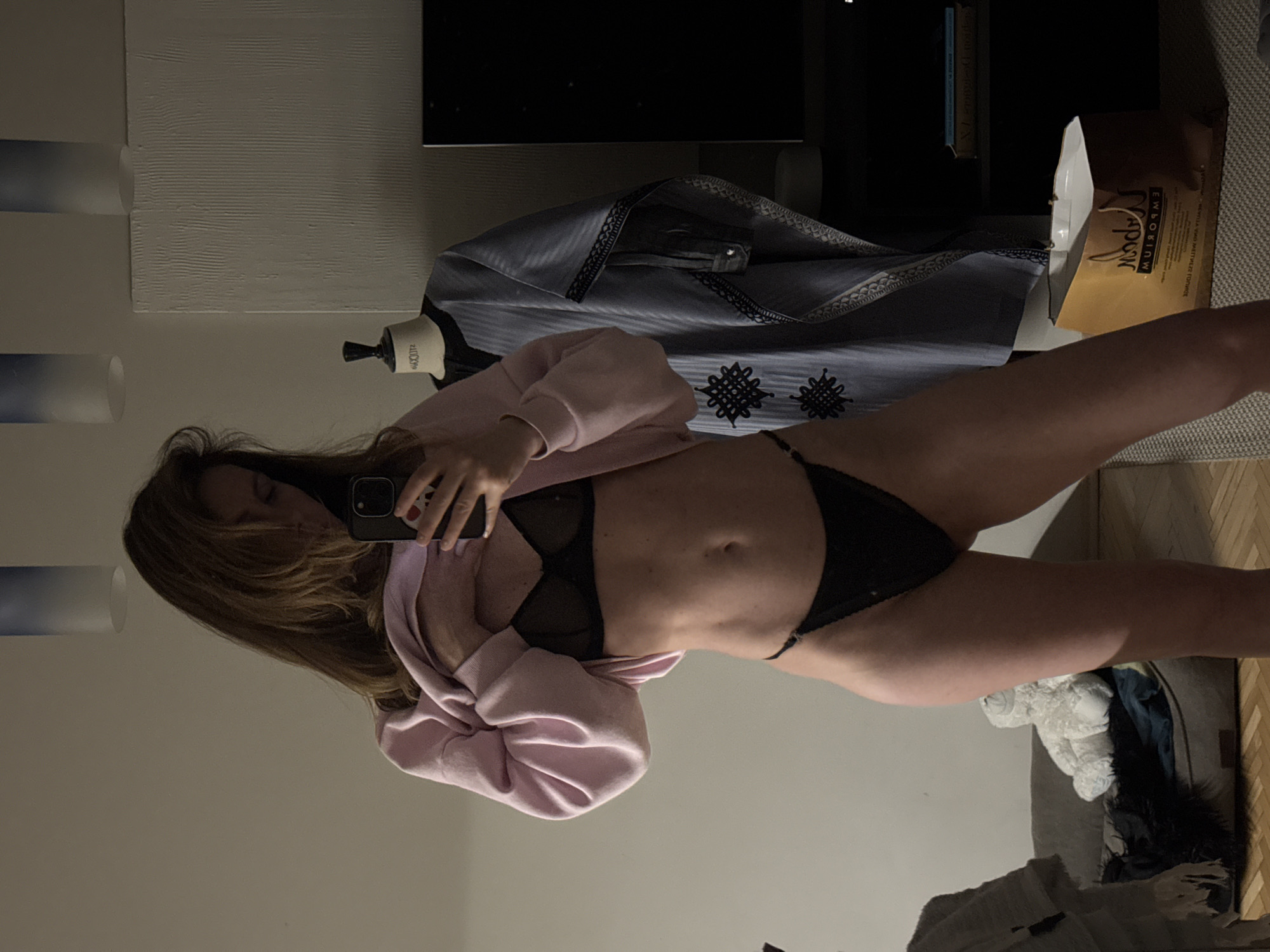 A woman wearing a black lingerie set and a pink sweater is taking a selfie in a mirror. Behind her are a dress form with a draped, patterned garment, a shopping bag, and a pet bed. The location cannot be determined from the image.