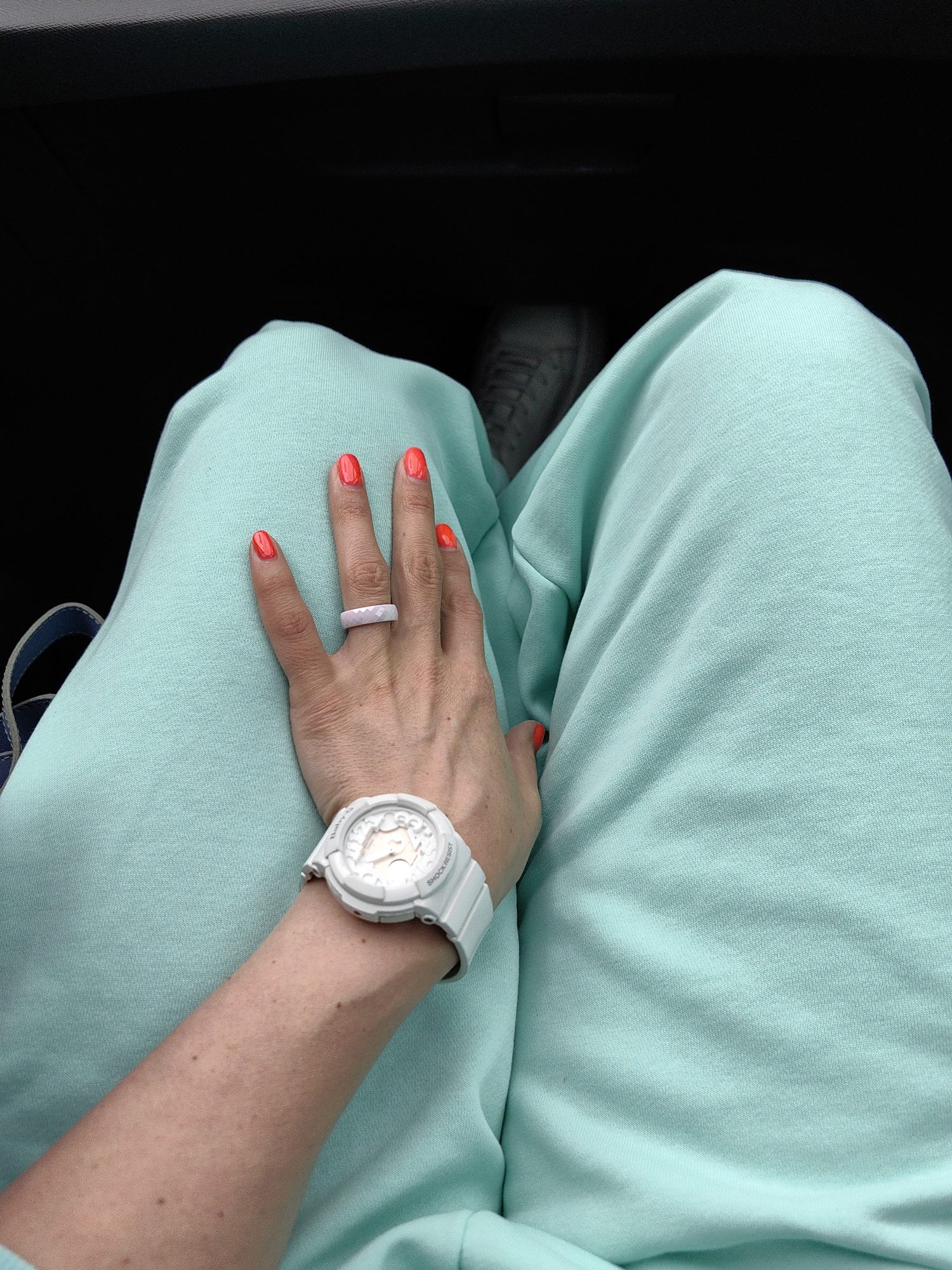 The image shows a person's legs and arm, wearing light teal sweatpants and a white watch. The person has bright orange nail polish on their fingernails and is wearing a light pink ring. A white shoe is visible on the left leg. The background is dark, suggesting an indoor or shaded environment. The image was taken in Putsčynski sel'ski Savet, Belarus.
