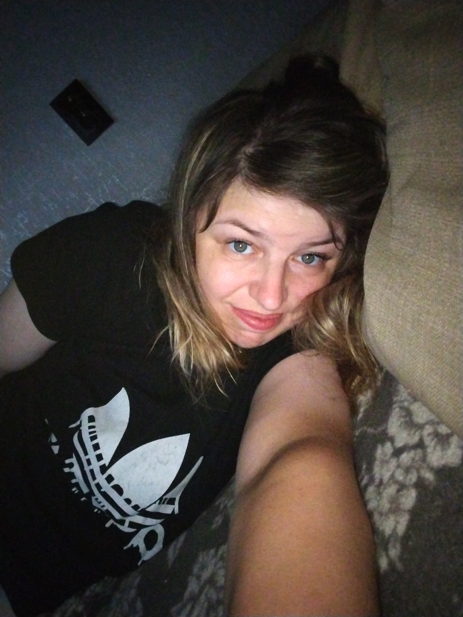 A woman with light brown hair and blue eyes lies on a couch, resting her head on her arm and looking at the camera. She is wearing a black t-shirt with a white Adidas logo. The background is dark, with a textured surface that appears to be a wall or floor and a portion of a couch. There is a small black rectangular object on the surface behind her head. The image appears to be a selfie taken indoors. The location in Yaroslavl, Russia, is not discernible from the image.