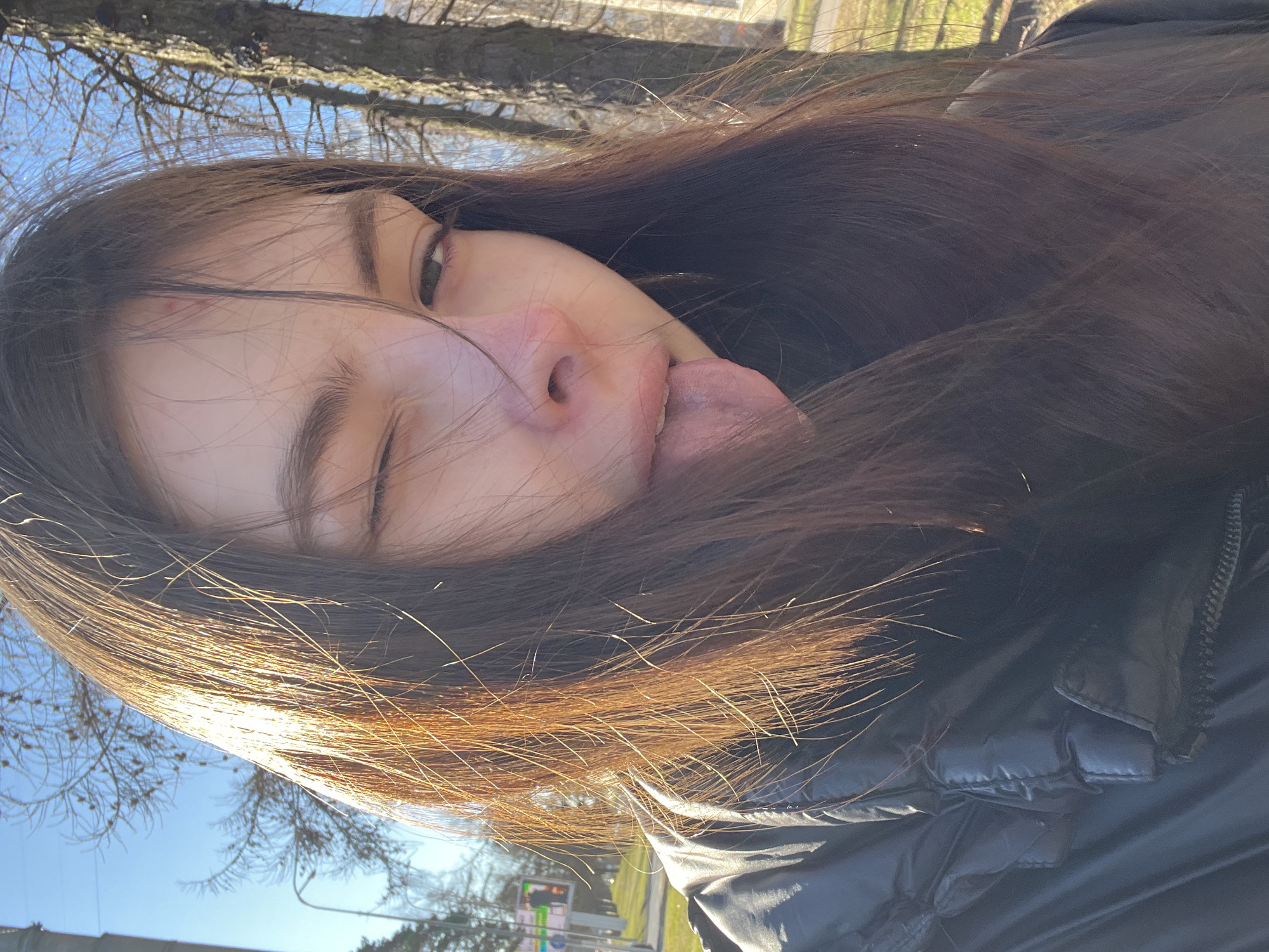 A young woman is pictured in a close-up selfie with a playful expression, sticking her tongue out and winking. Her dark hair is long and partially obscures her face. She is wearing a black jacket. The background is blurry, but trees are visible, and the sky is blue. The location is identified as Saint Petersburg, Russia. There are signs visible, but cannot be completely read.