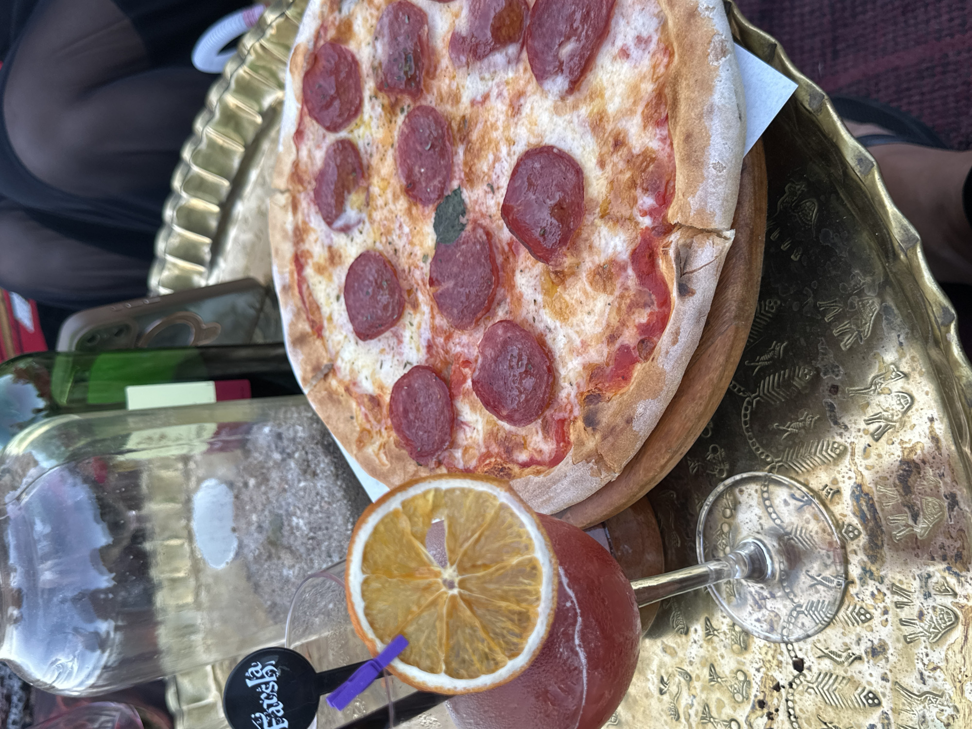 A high-angle shot captures a casual dining scene laid out on an ornate, aged golden/brass tray. The tray features intricate embossed patterns, including fish skeletons and other designs.
At the center, a small, personal-sized pepperoni pizza rests on a round wooden serving board. The pizza has a crispy crust, melted cheese, numerous pepperoni slices, and a visible green basil leaf. To the right, a glass containing a reddish beverage is garnished with a large dried orange slice and a purple stirrer. A black tag attached to the stirrer clearly displays the word "Eats".
On the left side of the tray, a large clear glass bottle, possibly a carafe, is partially visible, along with a smartphone encased in green. Further to the left, a portion of another bottle with a red label can be seen. In the upper left corner, part of a person's leg, dressed in sheer black fabric, is visible, indicating someone is seated nearby.
The scene is brightly lit, suggesting it is daytime. The arrangement suggests a relaxed meal or snack, possibly at an outdoor or semi-outdoor establishment in Sharm El Sheikh, Egypt.