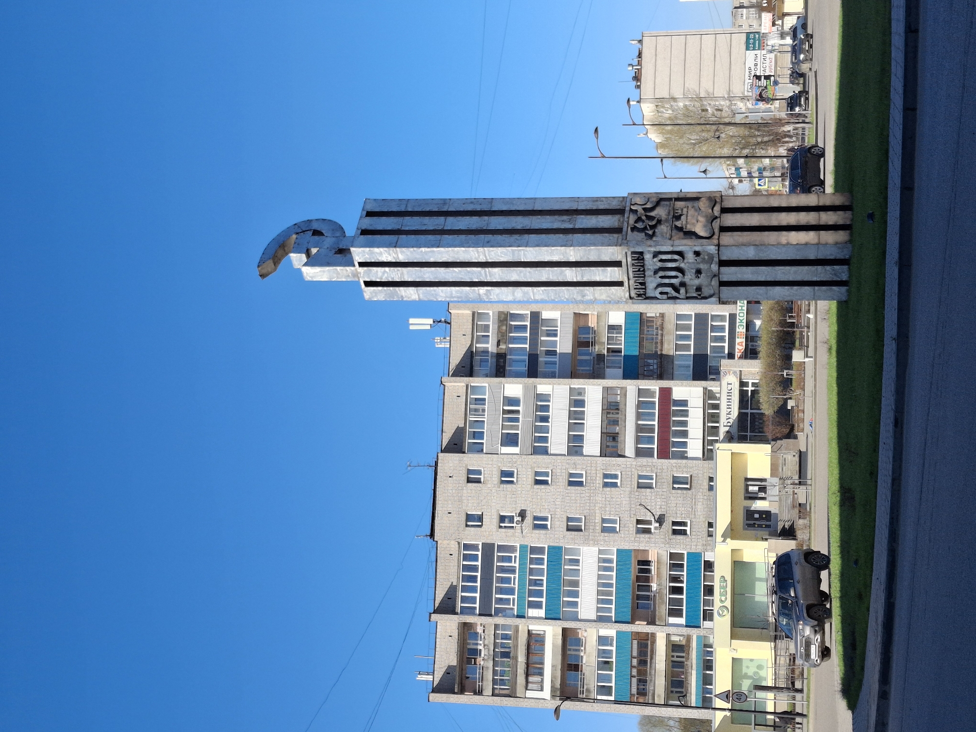 The image shows a monument in Sarapul, Russia, with a tall, metallic structure consisting of horizontal bars topped with abstract shapes. The monument's surface features inscribed text, including "200 Sarapulu." Behind the monument is a multi-story building with a mix of gray, blue, and white window frames. A commercial establishment with a "Sber" logo is visible, as well as a sign saying "Bukinust" and another with "Мир Кровли Анастил." A car is parked nearby. The sky is a clear blue, and overhead power lines are visible. The scene appears to be a sunny day in a city street.