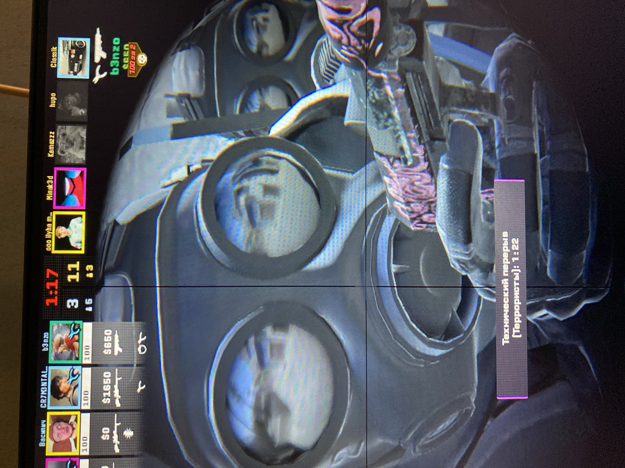 The image captures a first-person view within a video game, focusing on a character wearing a dark, tactical gas mask. The character is holding a weapon, possibly a rifle, which features a distinctive pink and purple patterned skin on its stock and grip. The reflections visible in the gas mask's eyepieces suggest an internal or enclosed game environment.
The left side of the screen displays the game's user interface, listing player names vertically. These include "Василич", "CR7M3NTAL", "b3nzo", "ooo ilyha m..", "Minak3d", "Kamazzz", "hupo", and "Classik". Adjacent to some names are player avatars, numerical values such as "$0", "$1650", and "$650" (likely in-game currency), and icons representing various weapons. A score display shows "1:17" (presumably a timer), "11", "3", and "5", alongside other stats like "100". One player, "b3nzo", is highlighted with additional details including "100 за 2" and a skull icon, indicating player performance.
On the right side of the screen, a prominent purple banner with white text displays a game message in Russian: "Технический перерыв (Террористы): 1:22". This translates to "Technical break (Terrorists): 1:22", indicating that the game, likely a tactical first-person shooter, is currently paused during a match. The image itself is a photograph of a screen, showing pixelation and potential light reflections.