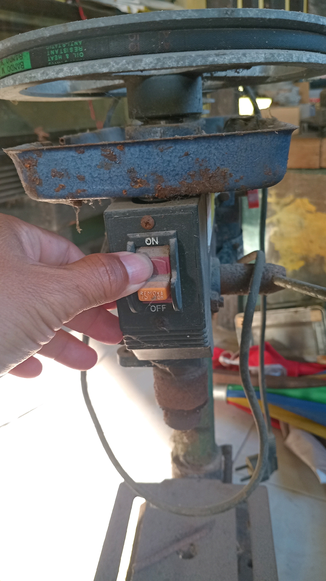 A close-up, eye-level shot shows a hand operating a red and orange toggle switch on a dusty, gray power mechanism, likely part of a drill press. The switch is positioned vertically and labeled "ON" at the top and "OFF" at the bottom, with "REMOVE TO LOCK" printed on the orange section. The hand, with visible skin texture and short fingernails, is pushing the switch upwards towards the "ON" position. The surrounding machinery appears old and weathered, with visible rust spots on a blue metal casing. A black belt runs across the top of the frame, connected to a pulley system. The background is blurred, showing hints of colorful fabrics and wooden structures, suggesting an outdoor or workshop environment. The image is taken during daylight, with bright light sources creating some overexposure. The setting is indoors, possibly a workshop or garage, in Randuagung, Indonesia, but this specific location information is not visually verifiable from the image itself.