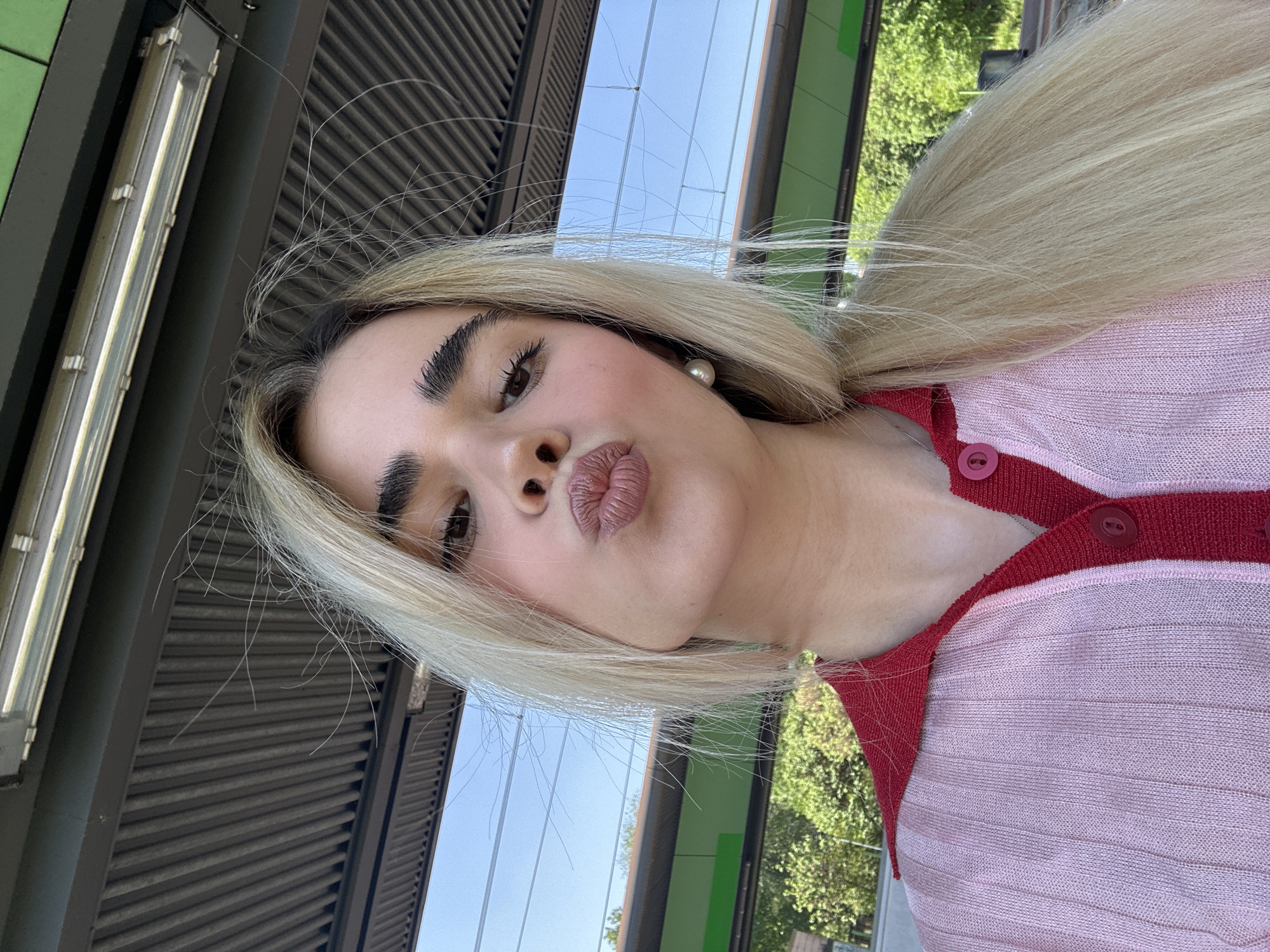 A young woman with fair skin, full lips, and blonde hair is centered in the image. She is wearing a pink and red striped button-up sweater. The woman is pouting slightly, facing the camera at an angle. The background shows an urban environment with a metal structure featuring horizontal slats and vertical green panels, visible through the gaps and possibly indicative of a public transit setting. Reflected in the slats above is a bright blue sky. The style and architecture suggest this may be a train or metro station. Given that the request specified Milan, Italy, it's reasonable to infer the location is a metro or train station within that city.