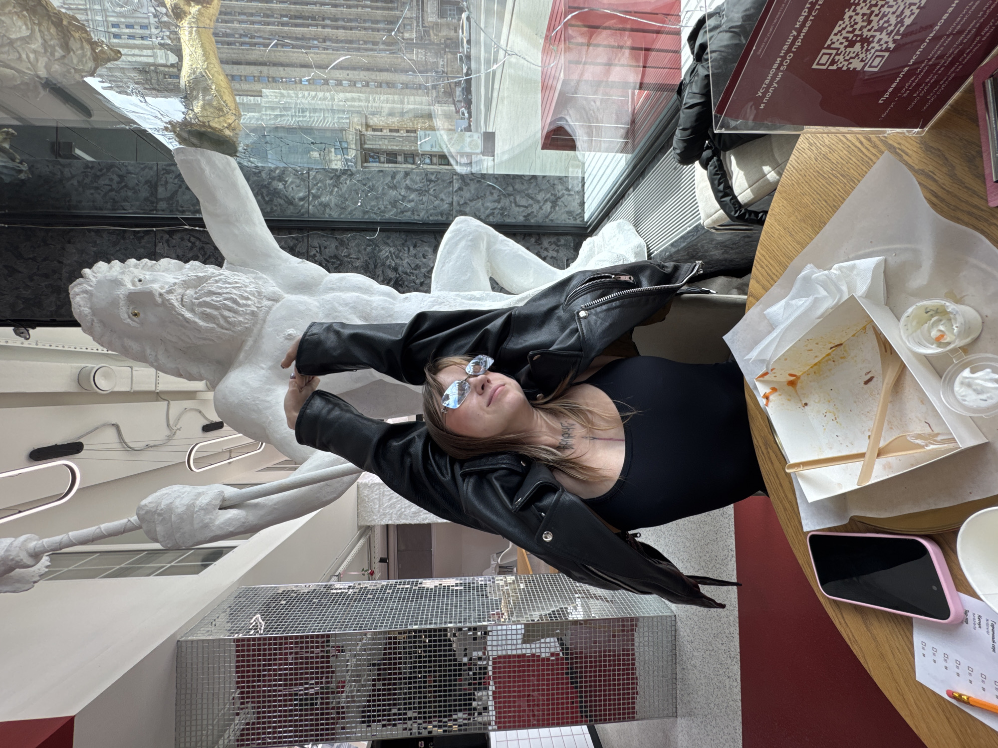 In this image, a person is lying down with their head tilted back, wearing a black leather jacket and sunglasses. They are positioned on a table next to discarded food containers and a cell phone with a pink case. The background features a large, white sculpture of a male figure, possibly classical in style, with its arm outstretched. Reflections in the glass behind the sculpture show an urban environment with buildings. To the right, a table holds a menu with Cyrillic text and a QR code, offering information about a loyalty program with bonuses and cashback based on spending. The overall setting appears to be an indoor cafe or restaurant with large windows. The lighting suggests it could be daytime.