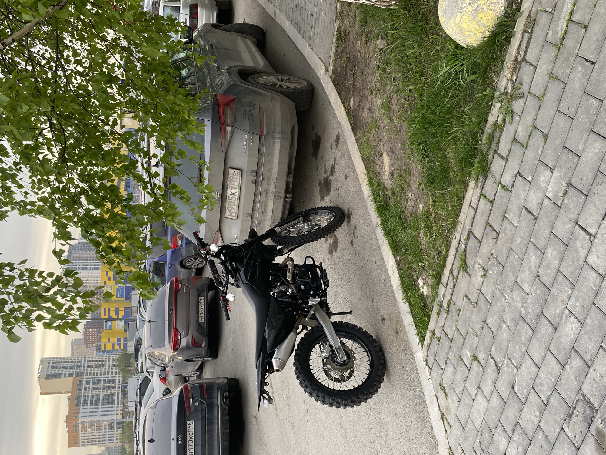 The image depicts a black dual-sport motorcycle parked near a row of parked cars on a city street in Yekaterinburg, Russia. The background features tall residential buildings. The cars include a gray Skoda Kodiaq with license plate M905KT196, a gray Kia with license plate M916XM196, and a black Kia with license plate H170XC196. The area appears to be a parking lot adjacent to a sidewalk paved with gray bricks. There is a grassy area between the sidewalk and the street. A tree with green leaves is visible in the upper left of the frame, partially obscuring the buildings in the background. The sky is overcast.
