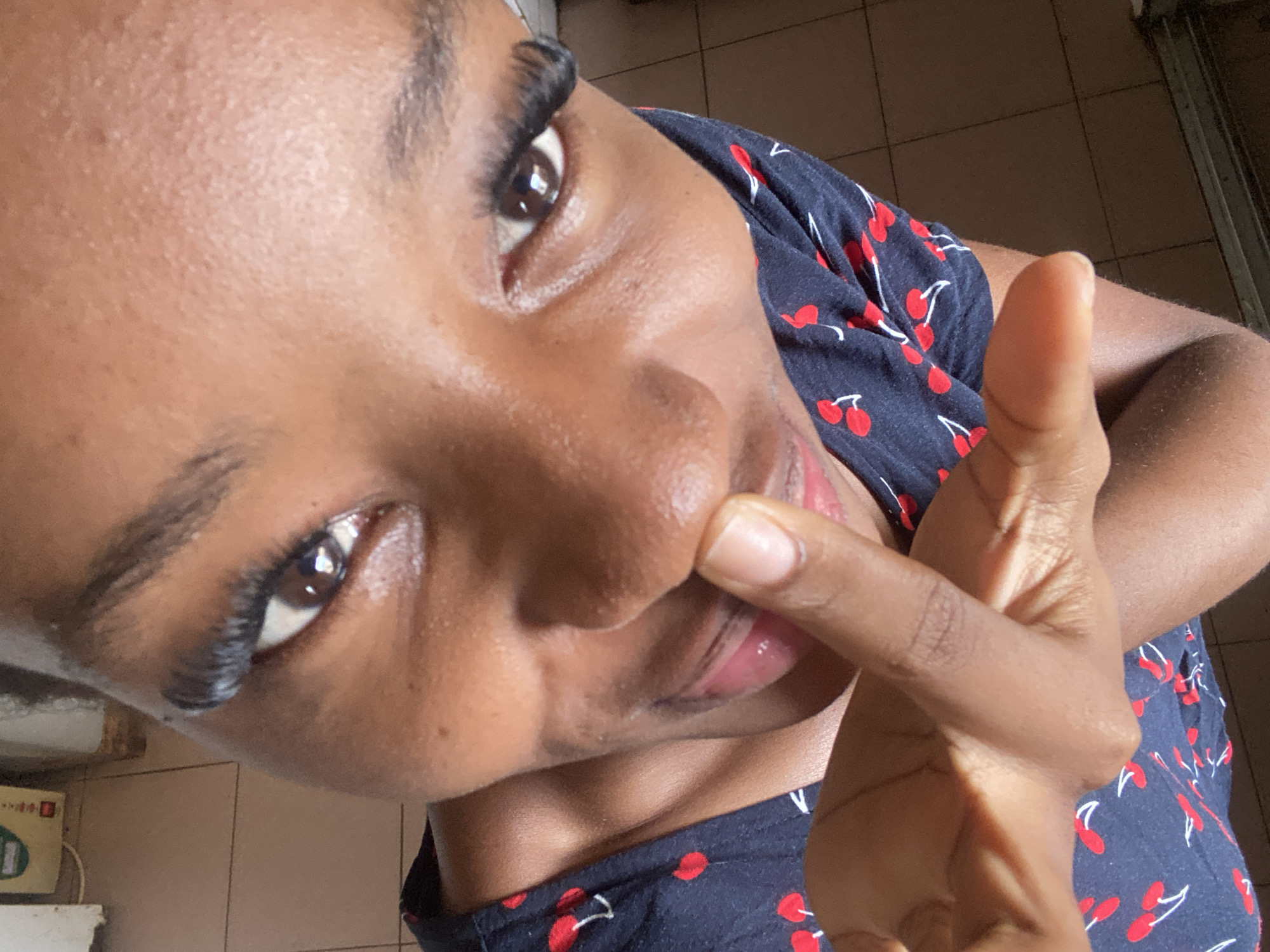 A high-angle, close-up portrait features an individual with a dark skin tone, looking directly into the camera. The subject's face occupies the majority of the frame. Both eyes are visible, characterized by dark irises and prominent, lengthy dark eyelashes. Dark, defined eyebrows are present above the eyes. The forehead and upper cheeks exhibit a subtle sheen. The right index finger of the subject is positioned against the philtrum area, partially obscuring the nose and upper lip. The mouth is partially visible, revealing a portion of the lower lip and some teeth, consistent with a slight smile. The individual is wearing a dark-colored garment, patterned with red cherries and white stems and leaves. The background shows a light brown tiled floor with visible grout lines in the lower right and upper left sections. A blurry, light-colored object and a faintly discernible green-and-white object are present in the upper left background. No visual elements within the image provide confirmation of the location being Jos, Nigeria.