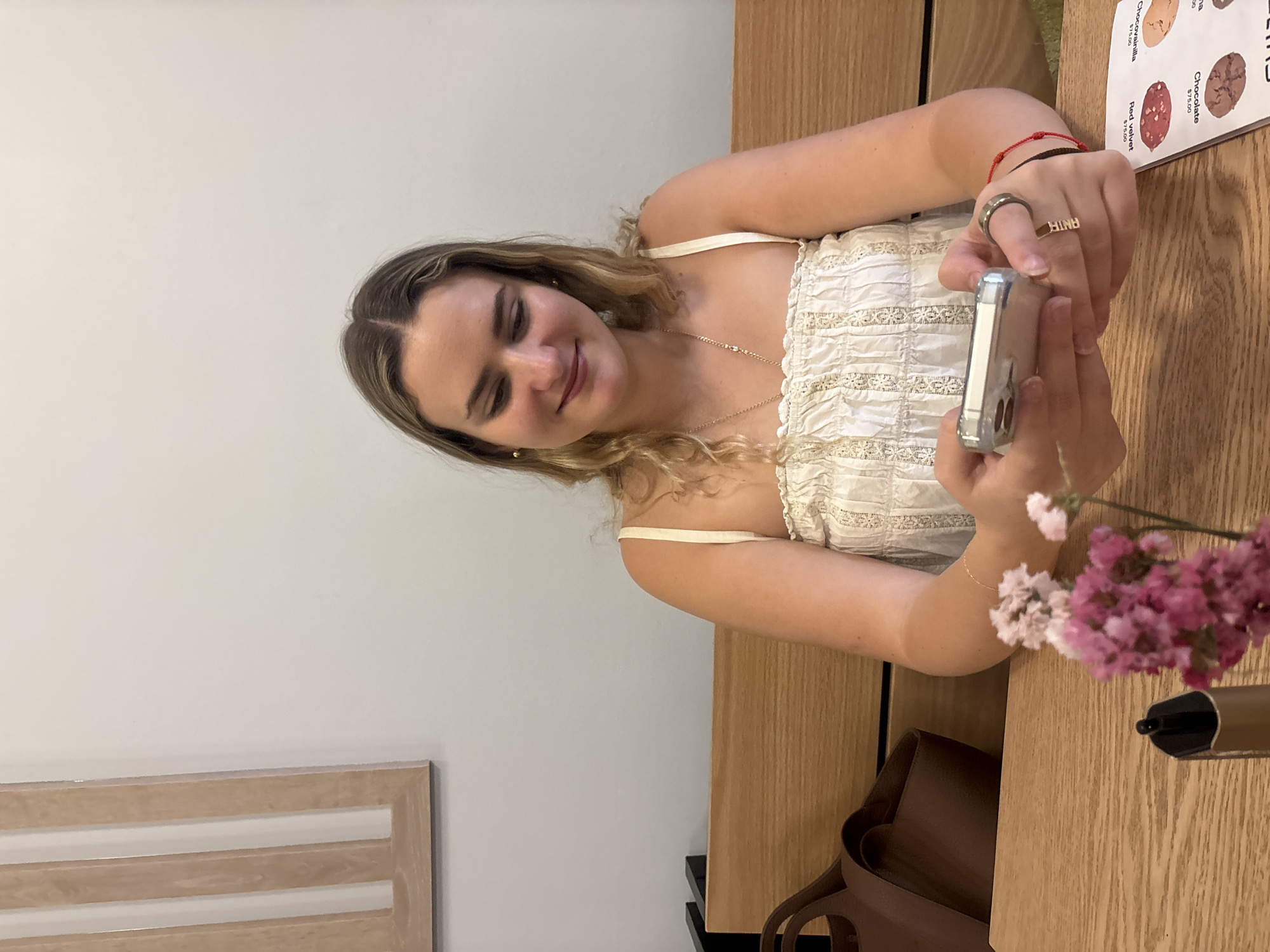 A woman is seated at a wooden table, holding a smartphone in her hands, possibly taking a picture. She has blonde hair and is wearing a light-colored, textured top with thin straps. She also has bracelets on her wrist. On the table, there is a menu with descriptions of food items and a small arrangement of pink flowers in a glass. A brown handbag is on the floor beside the table. The location is likely a restaurant in Puebla, Mexico.