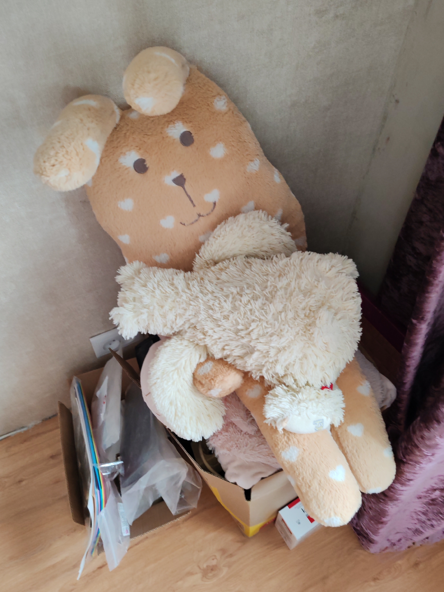 A tan, heart-patterned plush toy is resting in a cardboard box. A second, off-white plush toy is situated on top of the first. The box appears to be partially filled with assorted items, including what appear to be plastic-wrapped objects. The scene occurs indoors. The location is unknown and cannot be determined from the image.