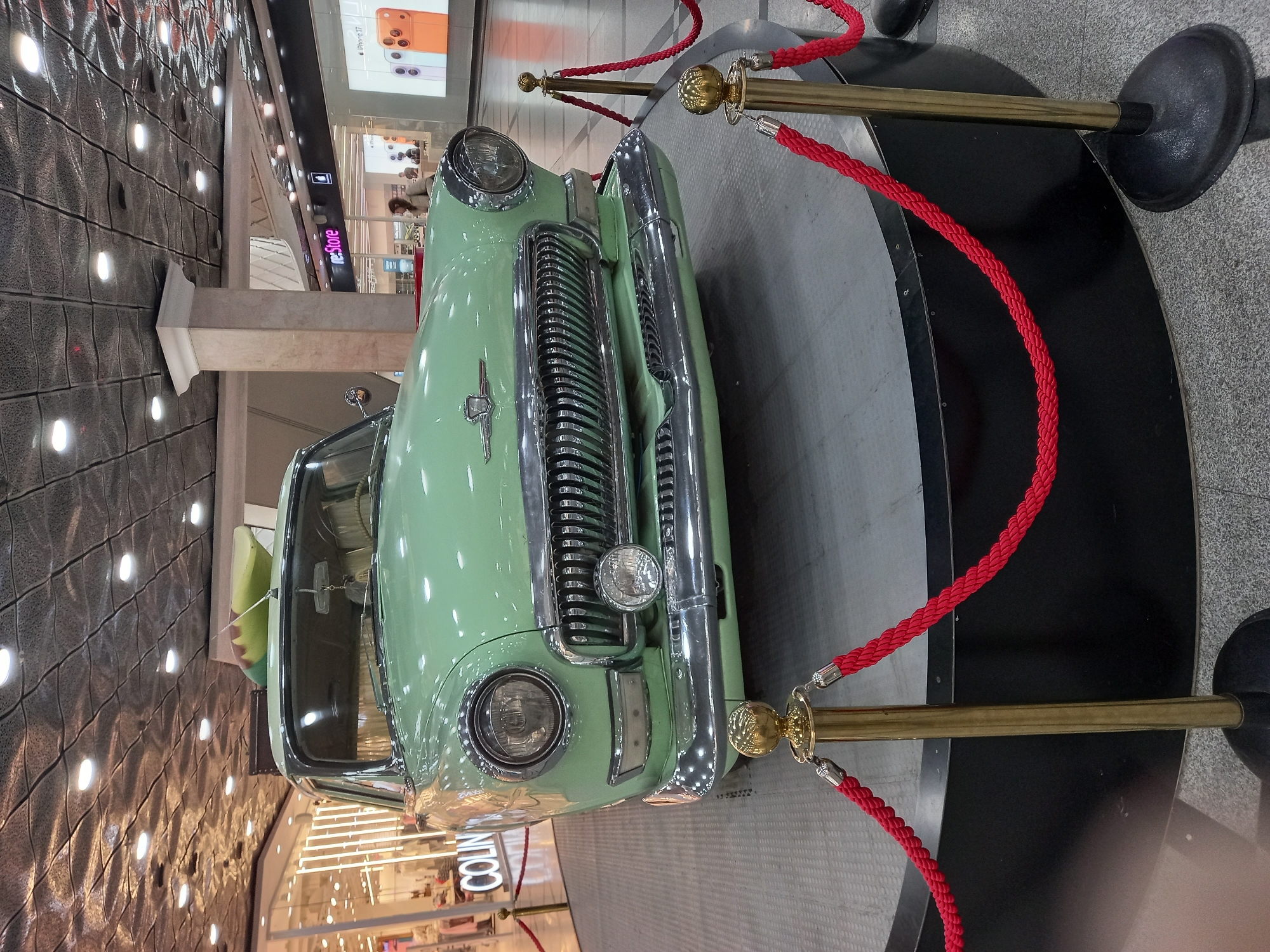 The image shows a vintage light green car displayed on a raised platform in what appears to be an indoor shopping center. The car has a chrome grille, round headlights, and a luggage rack on the roof with a green object. Red ropes are strung between gold stanchions, creating a barrier around the display. In the background, there are storefronts with signs such as "COLIN'S" and "re:Store," and posters for iPhones are visible. The ceiling has a textured pattern with recessed lighting. The presence of multiple retail establishments and the indoor setting suggest a commercial area within a city. The specific location of Yekaterinburg, Russia, is provided in the prompt but is not directly ascertainable from the visual information in the image itself, which lacks any overt Yekaterinburg-specific landmarks or signage. There are no discernible people actively interacting with the car or the display in the immediate foreground.