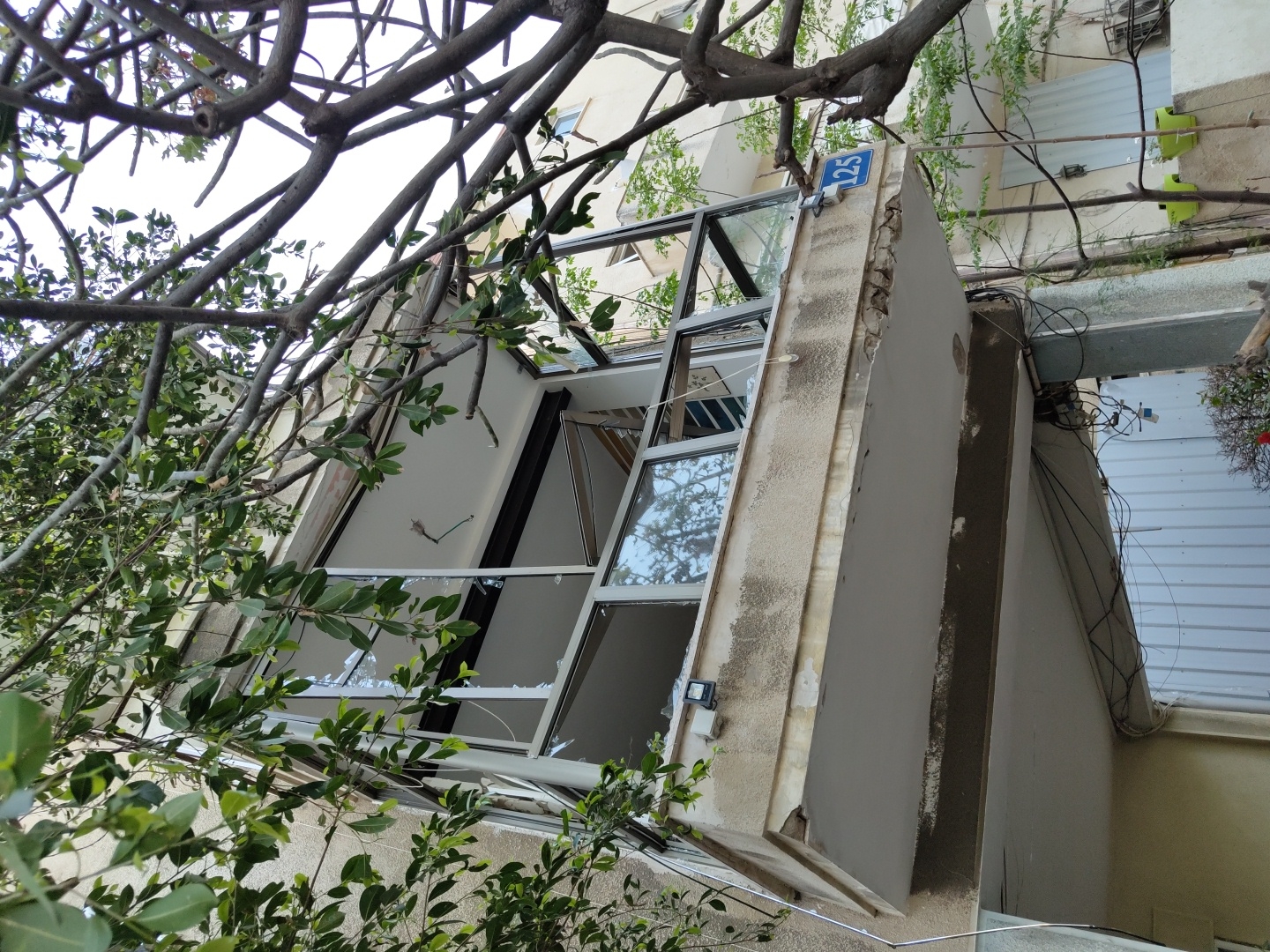 The image depicts a building with significant structural damage, possibly from a recent event. The window is severely damaged with broken glass and a displaced frame. A number "125" is visible on the building exterior. Green foliage is present in the foreground, partially obscuring the view of the building. A network of wires runs along the building facade. The scene appears to be urban, possibly in Tel Aviv, Israel, judging by the building style and architectural details. There are no people visible in the image.