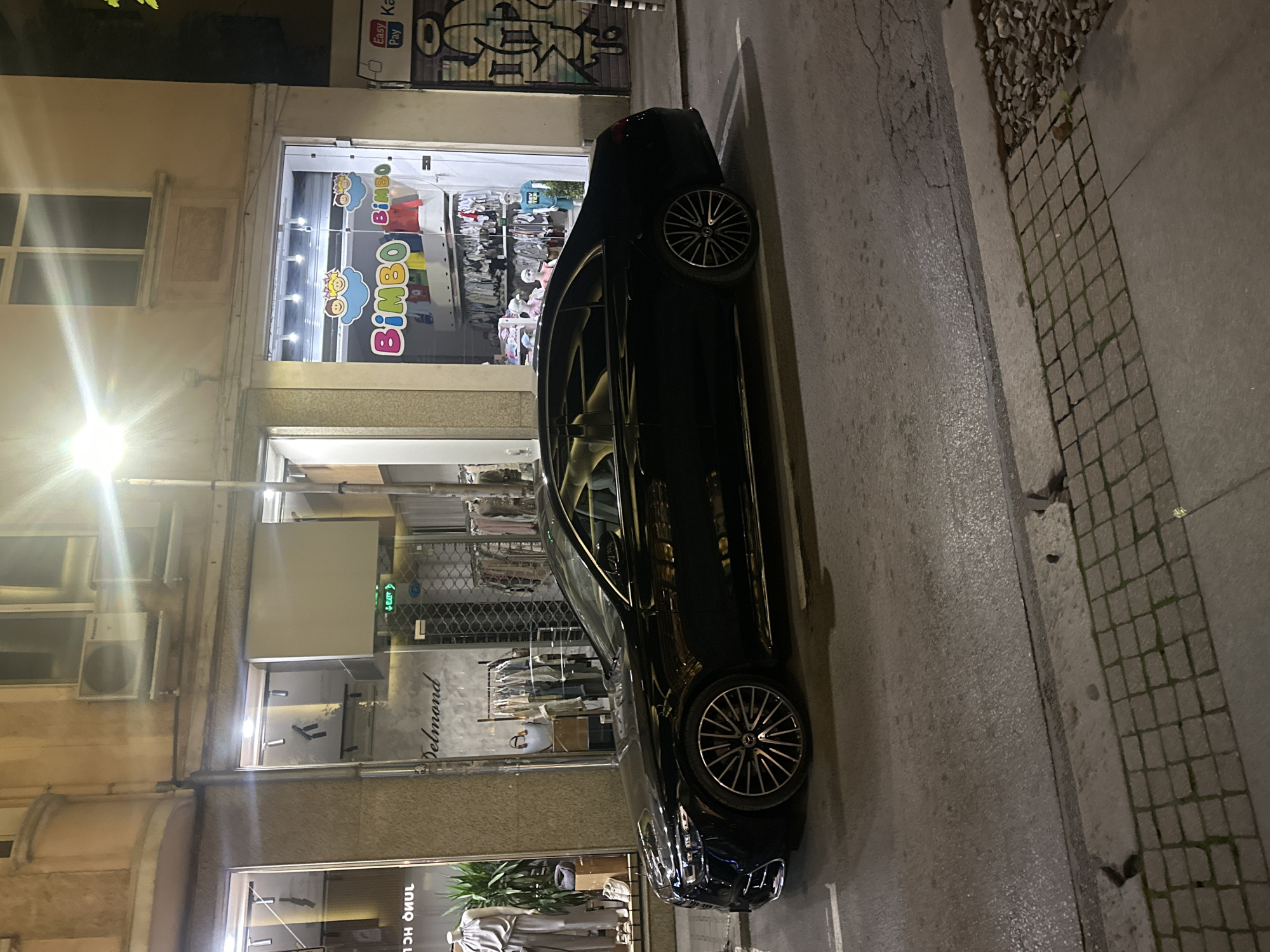 The image shows a dark-colored car parked on a street in Sofia, Bulgaria. The car is parked next to several storefronts, including a children's clothing store labeled "Bimbo", and a clothing store labeled "Delmond". The street is paved. The time of day appears to be night. The lighting is provided by street lights and storefront illumination. There are no people visible in the frame.