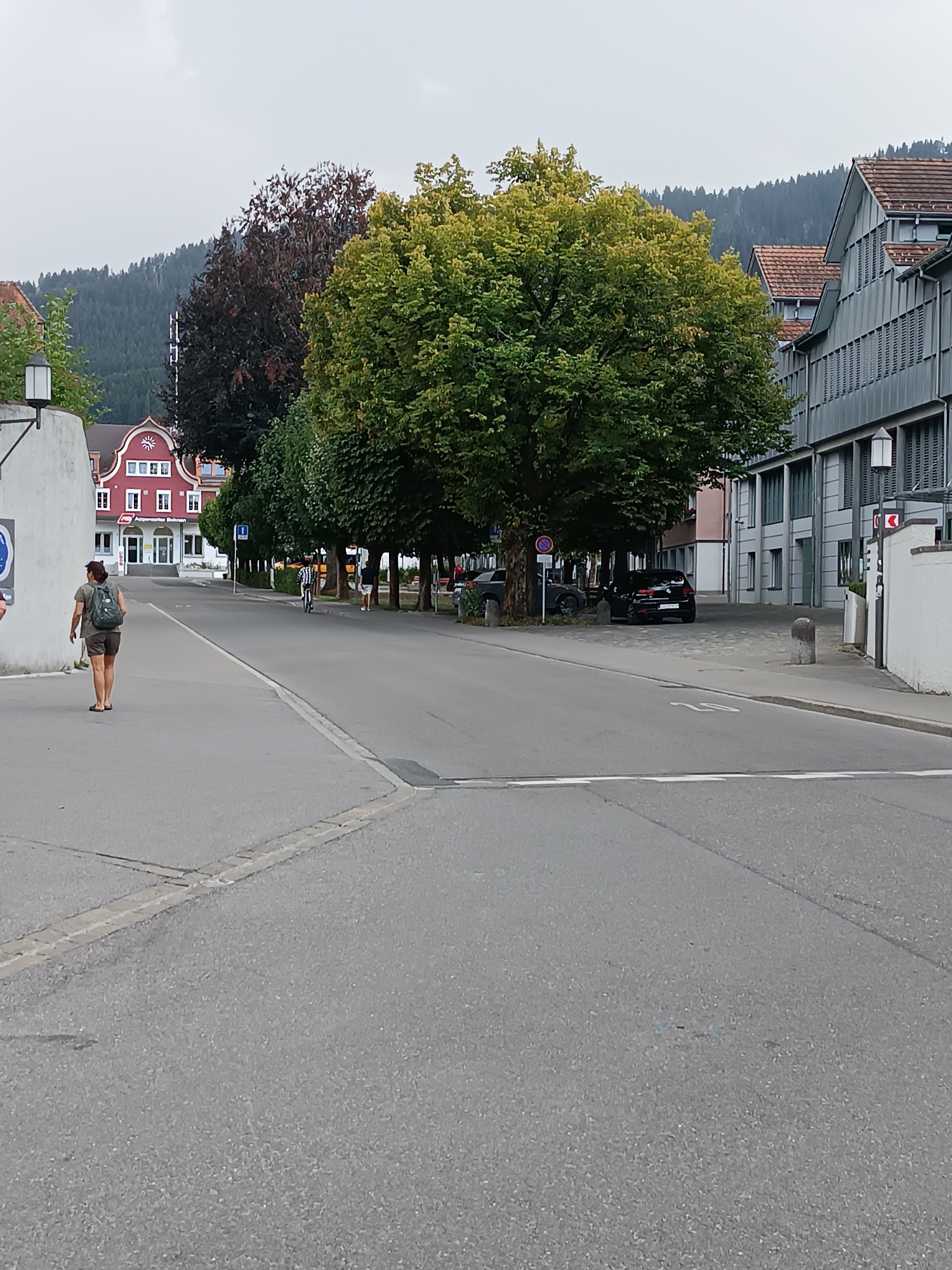 In Appenzell, Switzerland, a woman with a backpack walks on a paved sidewalk in front of a street. She is facing away from the camera, wearing a dark t-shirt and shorts. To her left, a white building with a black lamp is visible. Across the street, there are trees and several buildings, including a red building with white trim and a sign that reads "Appenzeller Bahnen." A black car is parked on the street. In the background, a forested hill rises against a cloudy sky.