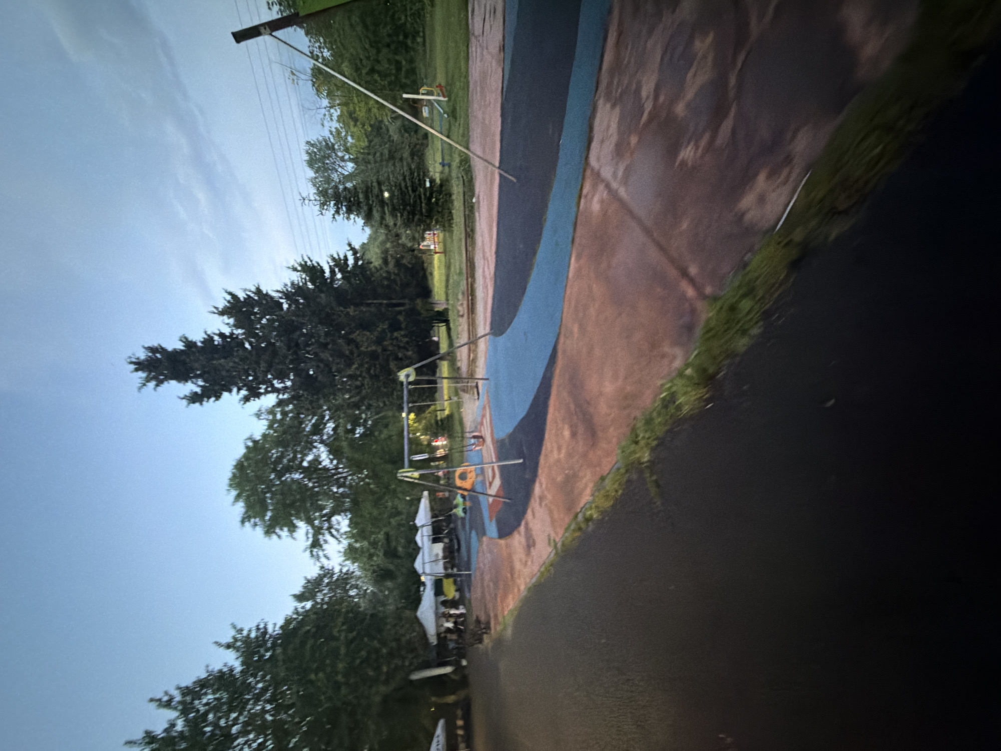 An outdoor scene in Burgas, Bulgaria, captured at dusk or early evening, shows a park or playground area. The image features a dark paved path in the foreground, separated by a strip of green grass from a larger, irregularly shaped paved area. This main area is distinctively colored with sections of brown, dark blue, and light blue, likely serving as a specialized surface for a playground.
In the midground, a swing set with a yellow seat is visible on the colorful pavement. Behind the swing set, several white pop-up tents are erected. Under and around these tents, numerous people are gathered, some standing and others seated, suggesting a community event, festival, or social gathering. The lighting under the tents appears somewhat artificial or reflective, contrasting with the fading natural light.
The background is dominated by lush, dark green trees, mostly conifers, framing the scene. A utility pole with wires extends into the sky on the upper right side of the original image. The sky, visible above the trees, is a soft light blue with faint cloud formations, indicative of a twilight hour. The ground in places appears damp or wet, reflecting the available light. No discernible text is visible in the image.