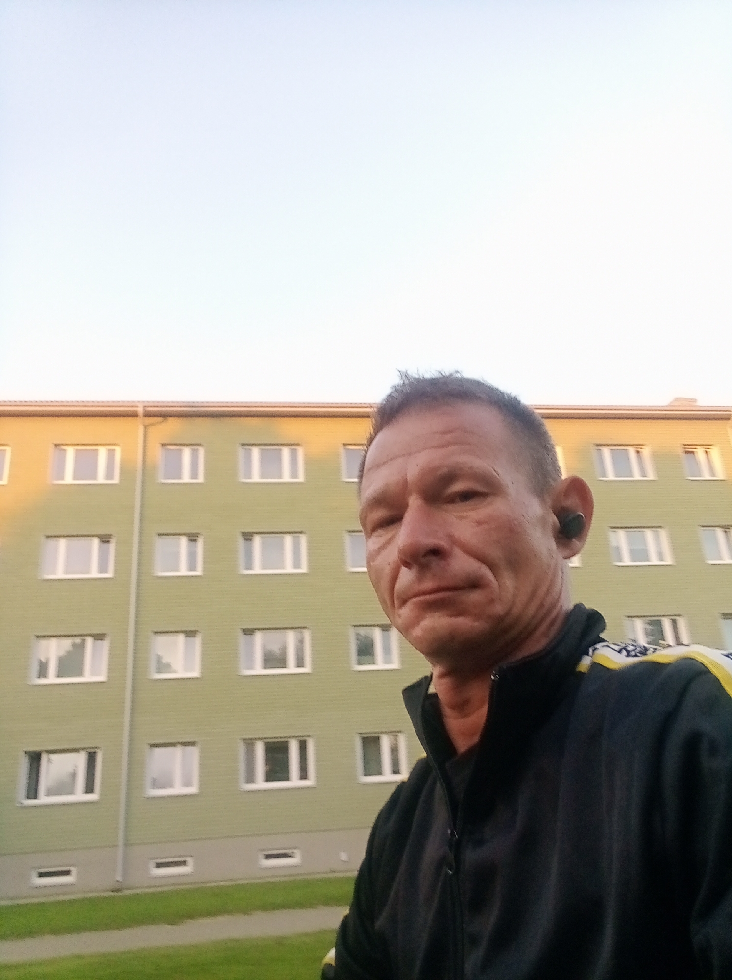 A man in his late 40s or early 50s with short, dark hair and a weathered face is taking a selfie outdoors. He is wearing a black zip-up jacket with yellow and white detailing on the shoulders. He has black earbuds in his ears. Behind him is a pale green, multi-story apartment building with numerous windows. The building appears to be in the city of Tapa, Estonia. The sky is clear and light blue. A grassy area is visible in the foreground.
