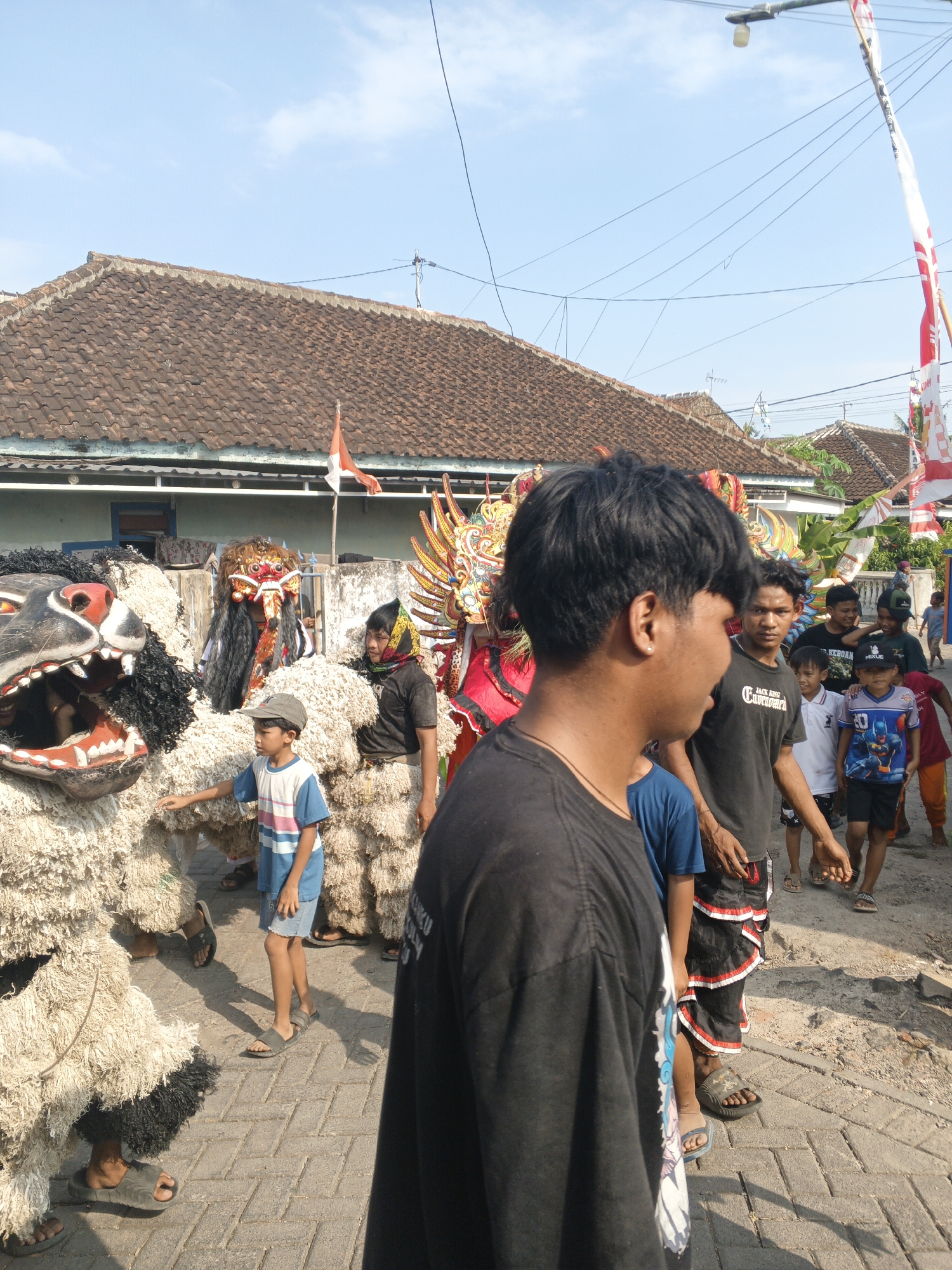 In Singolatren, Indonesia, an outdoor procession is underway on a sunny day. The scene is dominated by a large, elaborate costume resembling a mythical creature, possibly a Barong, made of light-colored woven material and black fur accents. Its open mouth reveals painted teeth and tongue. In front of this creature, a young boy wearing a cap, a striped t-shirt, and denim shorts is standing. He is looking towards the creature and has his left hand extended.
Behind the main creature, another costumed performer is visible, this one featuring a headdress with colorful feathers and elaborate facial markings. Further back, a group of people, including several young men and boys, are walking or standing. Some are looking towards the procession. The background features traditional Indonesian houses with tiled roofs, a clear blue sky, and utility poles with wires. Red and white flags, characteristic of Indonesian national flags, are displayed on poles, suggesting a festive or national event. The ground is paved with interlocking bricks. The overall atmosphere appears to be one of cultural celebration or parade.