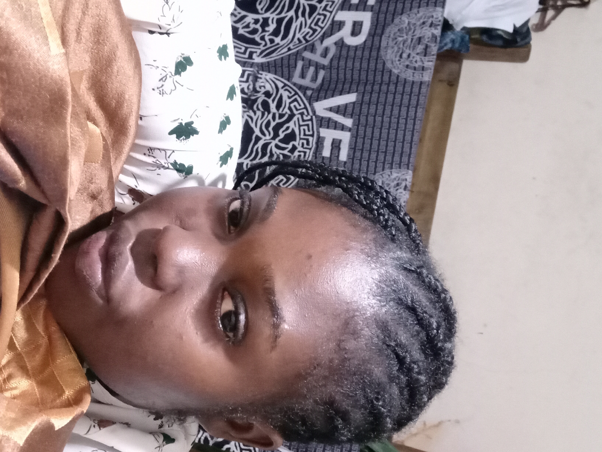 A close-up, low-angle selfie captures a young Black woman with dark skin and braided hair. She is looking directly at the camera, with her face occupying most of the frame. Her right eye is visible, along with her nose and lips. She is wearing a white top with a floral pattern and a shimmering gold fabric is draped over her chest and lap. Behind her, a patterned fabric with the letters "R VE" and a circular design is visible, suggesting she is in a domestic environment, possibly in Ejule, Nigeria. The background is a plain, light-colored wall. The lighting is soft, with shadows cast on her face, indicating an indoor setting.
