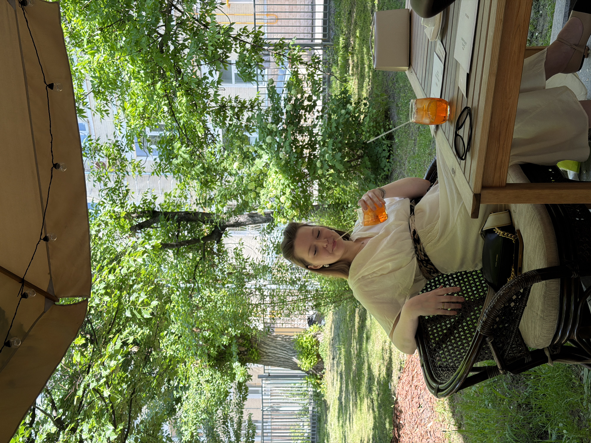 The image shows a woman seated at a table outdoors. The setting appears to be in a green, outdoor dining area, with trees visible in the background. The table holds a drink, possibly an Aperol spritz, and a pair of sunglasses. The woman is holding a drink in one hand and has a handbag on her lap. The scene is in Moscow, Russia.