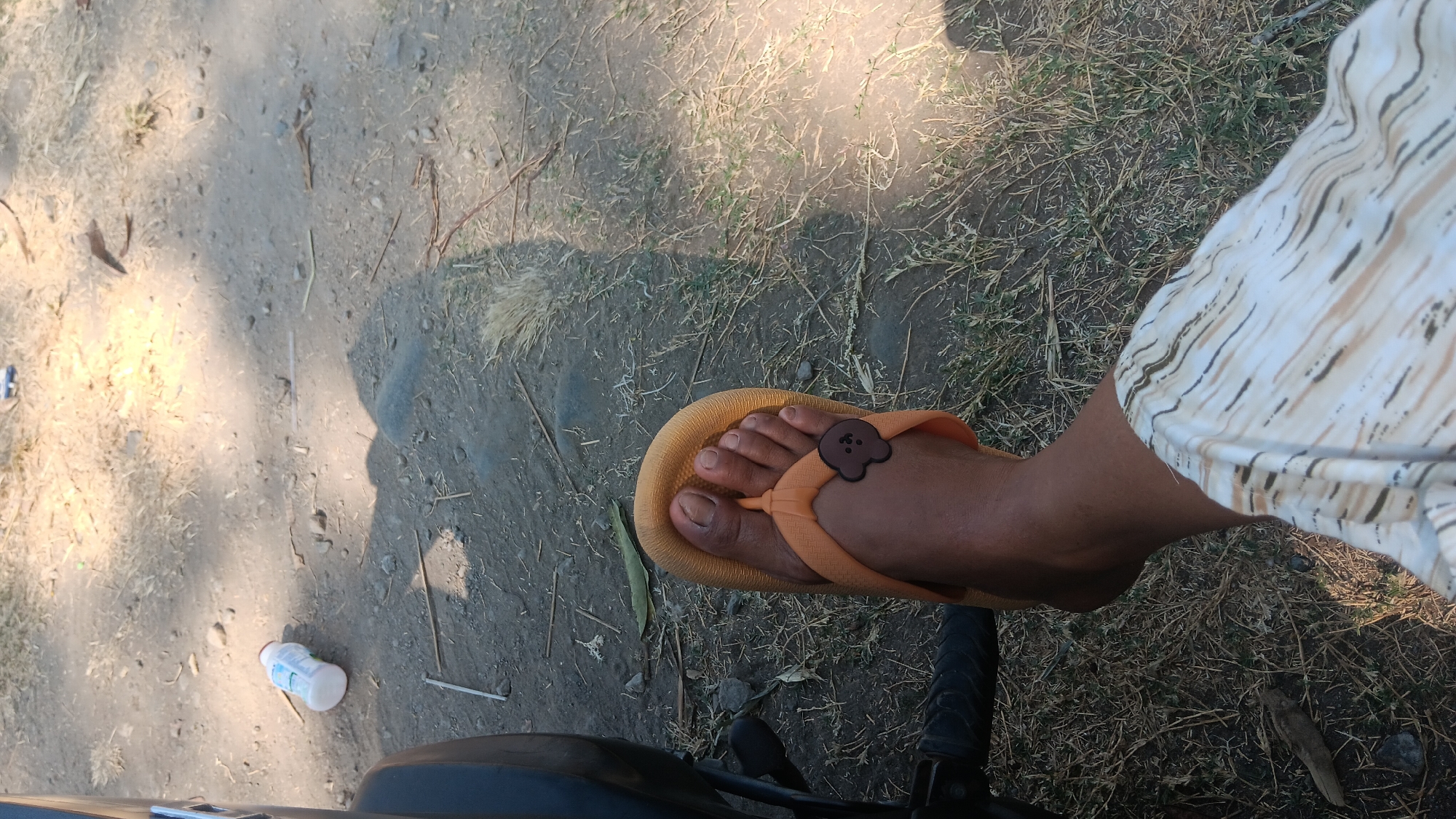 A low-angle, close-up shot captures a person's right foot, adorned in an orange flip-flop with a brown bear charm, resting on dry grass and dirt. The background reveals the ground, scattered with small stones and dry vegetation, with a partially visible black motorcycle on the left. A white bottle with a blue label lies on the ground in the upper left quadrant. The scene is illuminated by sunlight, casting shadows on the ground. The image was taken in Gemenggeng, Indonesia.