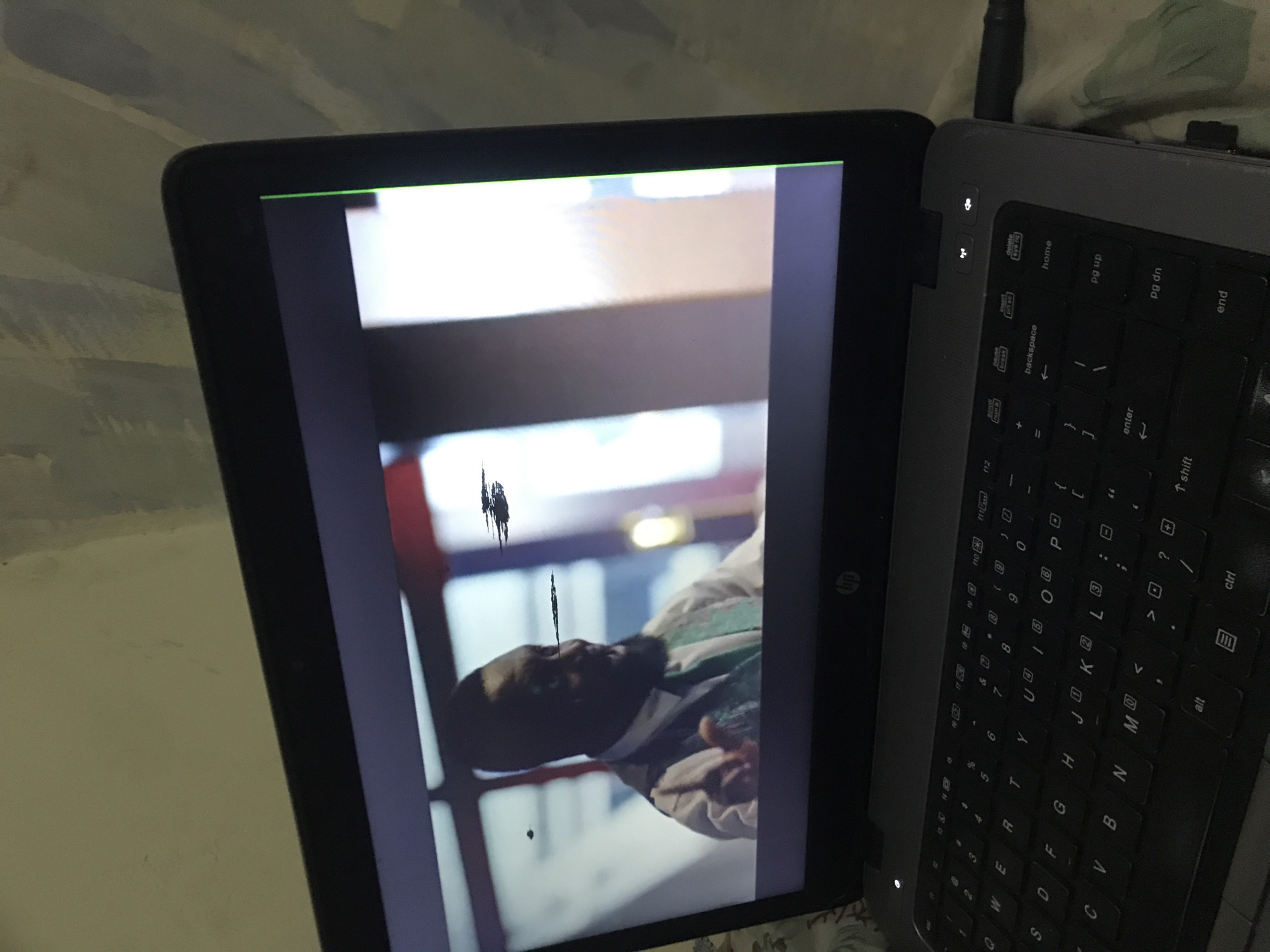 An indoor scene in Rafin Par, Nigeria, features an open HP laptop displaying a video. The laptop has a dark gray chassis and a black QWERTY keyboard with white lettering. A small white power indicator light is illuminated on the left side above the keyboard. A black cable is connected to the right side of the laptop. The laptop screen shows a video featuring a dark-skinned man with a beard, wearing a white shirt, a dark tie, and a green patterned vest, gesturing with his right hand. The video display on the laptop screen is marred by multiple horizontal dark smudges or artifacts and a prominent continuous vertical green line on the far right edge. The laptop rests on a light-colored, possibly patterned soft surface. Behind the laptop, a light beige or off-white wall with subtle textural variations is visible.