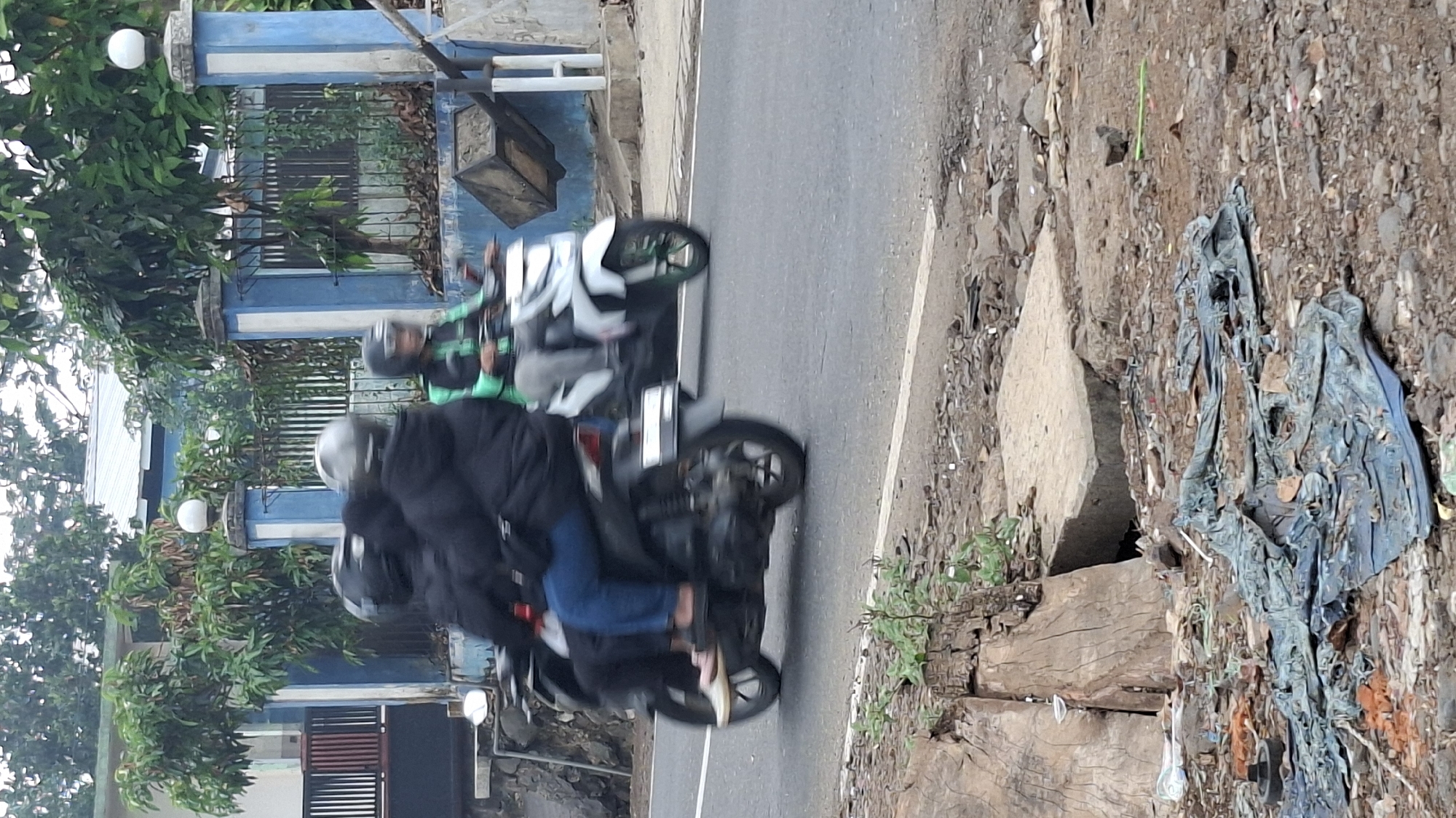 The image depicts a street scene in Cilaku, Indonesia, with two motorcycles on a paved road. The motorcycle in the foreground, on the left, is black and carries three individuals. The driver is wearing a black jacket and helmet, and the two passengers behind are also in dark clothing and helmets. The passenger in the middle is wearing blue jeans, and the person furthest back is partially obscured. The motorcycle itself has a visible license plate.
To the right, a white motorcycle is also in motion, carrying two individuals. The driver is wearing a green jacket and helmet, and the passenger behind is wearing a dark jacket and helmet. This motorcycle also has green accents on its wheels.
The background shows a building with a blue and white fence. Trees and foliage are visible behind the fence. The foreground of the image features a rough, unpaved area with debris, including torn fabric, rocks, and what appears to be discarded plastic. A concrete structure is also present in this area, possibly part of a drainage system. The overall lighting suggests an overcast day.