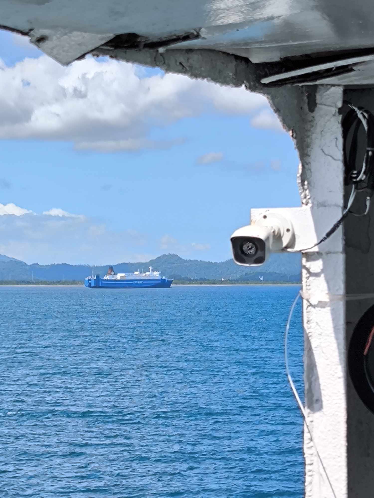 The image captures a vibrant daytime scene overlooking a calm blue sea, with a large blue ferry prominently featured in the middle ground. The photo is taken from a partially enclosed vantage point, with an overhead structure and a rugged white column or wall framing the right side and top of the shot.
The main subject is a blue and white ferry, sailing left-to-right across the frame. It has the text "CITY LINE" clearly visible in white letters along its hull. The ferry appears to be a passenger and/or cargo vessel, with multiple decks and a distinctive dark chimney or funnel at the rear. In the far distance, green, undulating mountains or hills line the horizon under a bright blue sky dotted with scattered white cumulus clouds.
In the immediate foreground, a white, cylindrical security camera is mounted on the rough, off-white structure to the right. Wires, one white and one black, are connected to the camera and run along the surface of the structure. The water in the foreground and mid-ground is a rich, deep blue with subtle ripples, indicating gentle conditions. The overall impression is of a sunny, clear day in a coastal or island setting. No people are visible.
