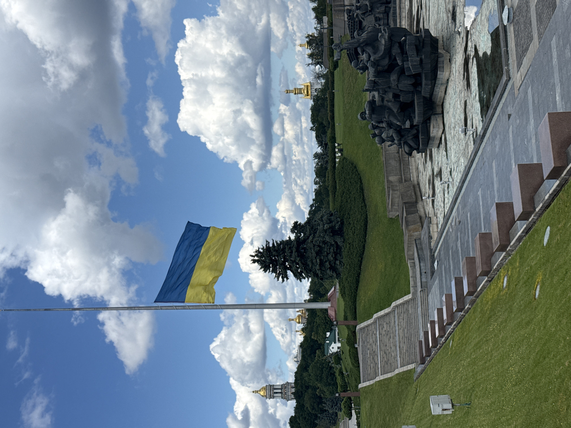 The image depicts the Eternal Glory Park in Kyiv, Ukraine. A tall flagpole stands prominently in the center, flying the Ukrainian flag at half-mast under a partly cloudy sky. To the left, a grand staircase leads up a grassy hill, flanked by granite steps. In the background, the golden domes of the Kyiv Pechersk Lavra are visible amongst lush green trees. In the foreground, a large, dark bronze sculpture depicting soldiers in battle is situated in a dry fountain area. The park setting features manicured lawns, trees, and pathways.