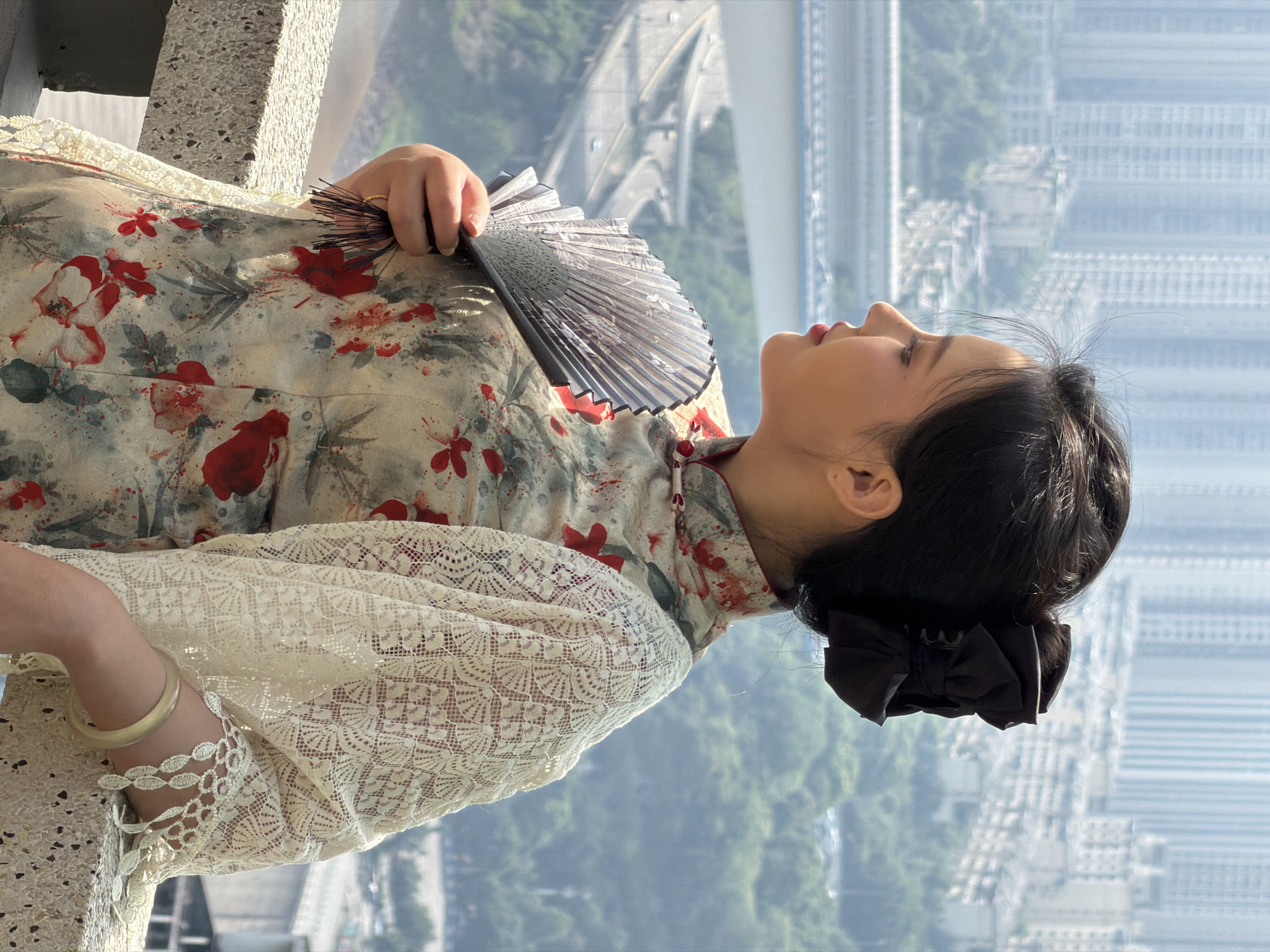 A young East Asian woman with dark hair styled in a bun and adorned with a black bow stands outdoors, her profile visible as she gazes to her left. She wears a traditional Qipao dress with a floral pattern and a cream-colored lace shawl. In her right hand, she holds a black fan. She is leaning on a stone railing, which appears to be part of a balcony or observation deck. The background is a cityscape with numerous tall buildings, suggesting an urban environment. Below the buildings, a multi-layered highway interchange is visible, with cars on its various levels. Green trees and a body of water can also be seen, indicating the presence of nature within the city. The lighting suggests it is daytime, possibly late morning or afternoon, with the sun casting shadows. The overall scene conveys a sense of serene contemplation against a bustling urban backdrop. The presence of the highway interchange and dense urban development strongly suggests the location is Yuzhong District, China, known for its characteristic mountainous terrain and intricate urban infrastructure.