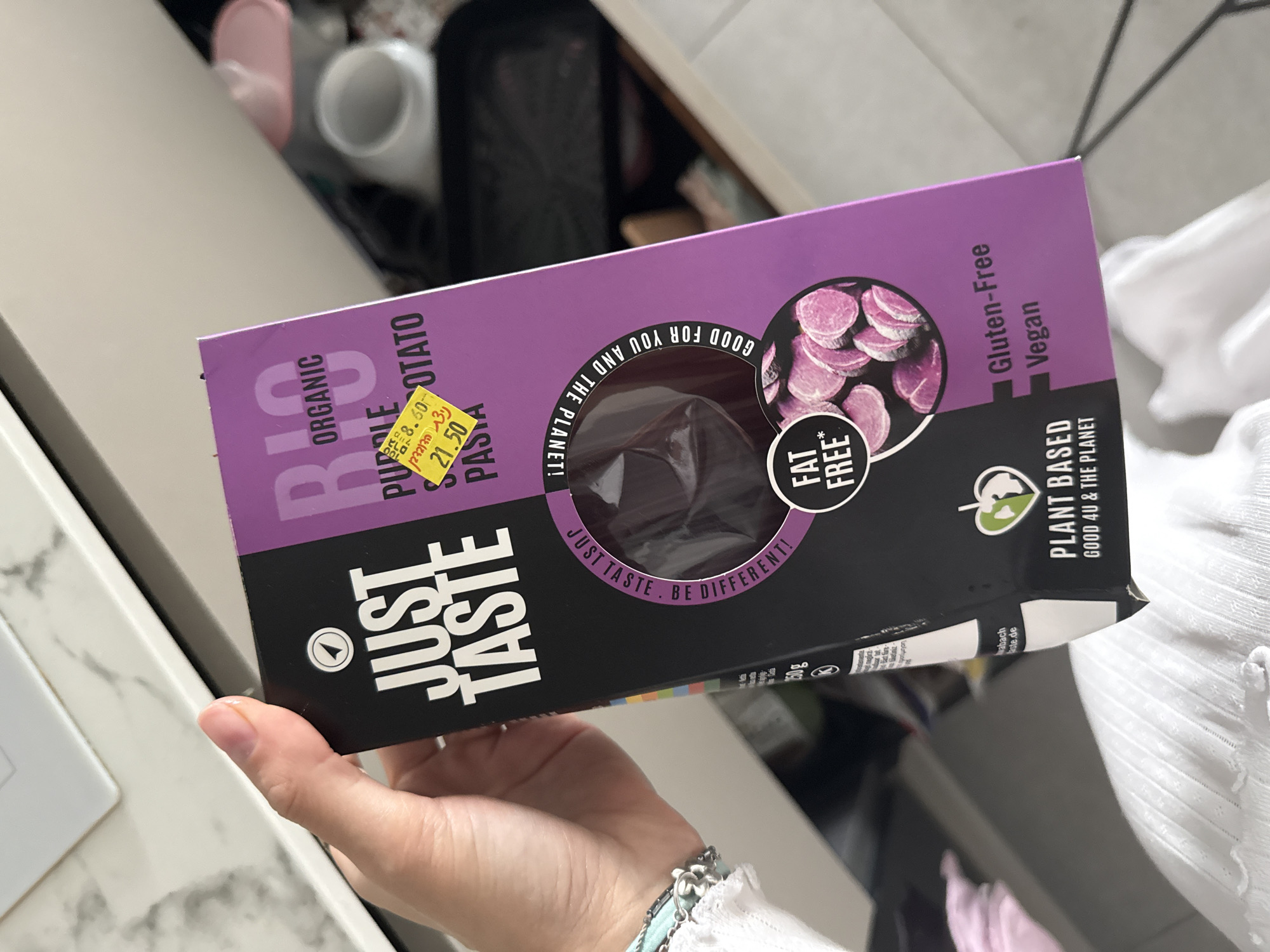 A hand is holding a purple and black box of "Just Taste" organic purple potato pasta. The box has a yellow clearance sticker on it. The background is a cluttered kitchen. It is not possible to confirm the location of this scene as Tel-Aviv, Israel.