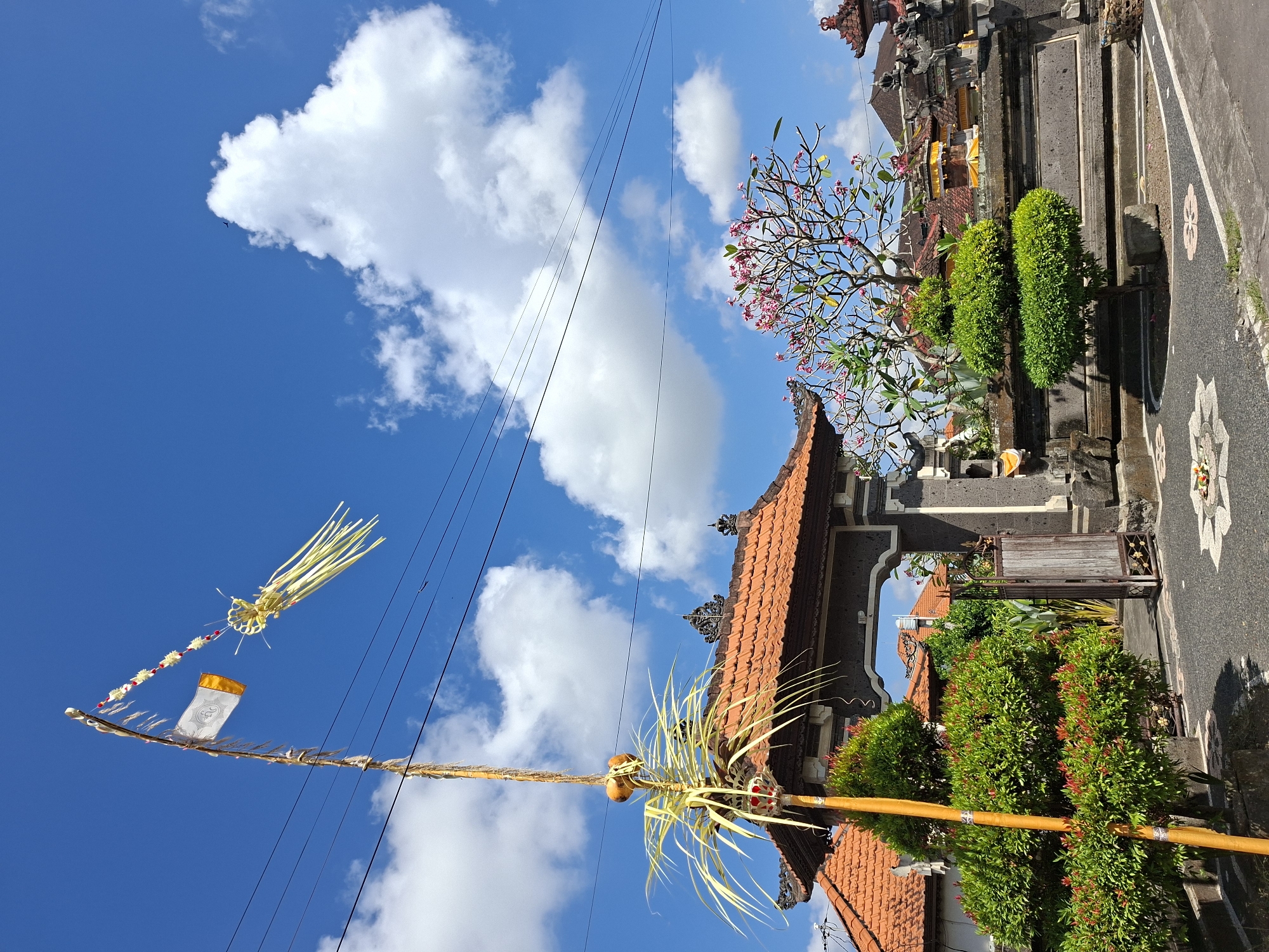 A tall, slender bamboo pole, adorned with yellow and white woven decorations and a small white and orange flag, stands in the foreground of a residential area in Blahbatuh, Indonesia. The pole is positioned to the left of a dark stone archway, which leads to a courtyard. The archway is flanked by lush green bushes and a weathered wooden gate. Behind the archway, the roofs of traditional Balinese houses, covered in orange tiles, are visible. A flowering tree with pink blossoms adds a touch of color to the scene, and intricately carved stone walls line the perimeter of the property. The sky is a vibrant blue, dotted with fluffy white clouds, and power lines stretch across the upper portion of the frame. The ground is paved with dark stones and decorated with floral patterns. The overall impression is one of cultural richness and a serene, sunny day.