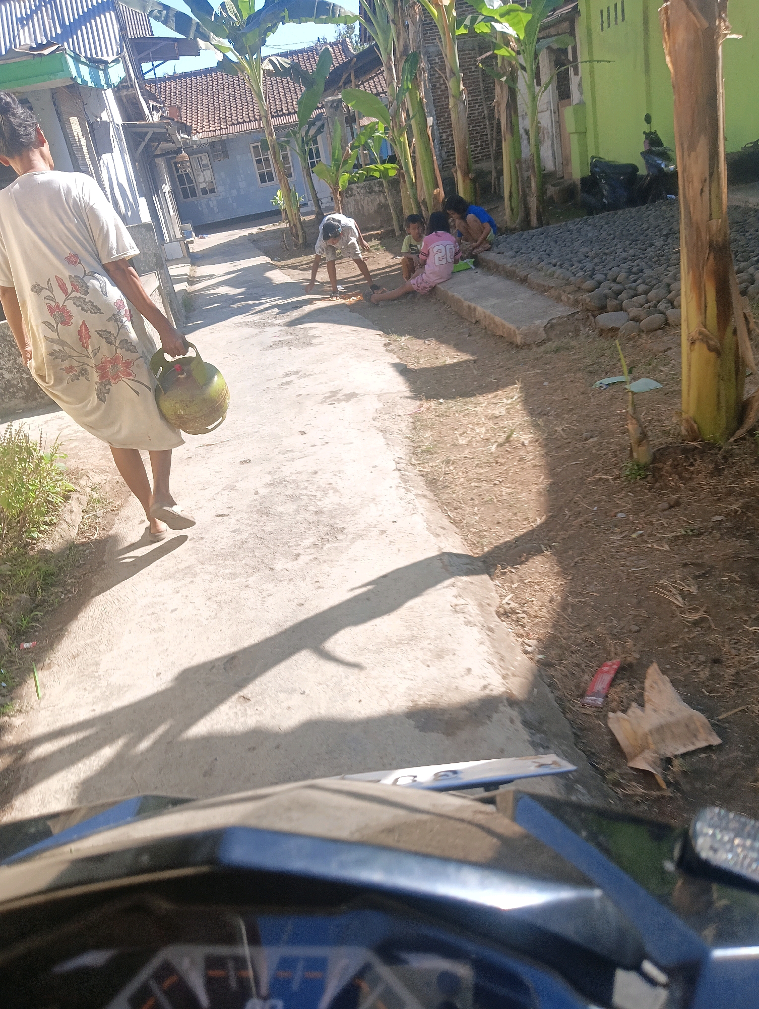The image depicts a sun-drenched outdoor scene in a residential area, likely in Sokaraja, Indonesia. In the foreground, the handlebars and dashboard of a motorcycle are visible, suggesting the photo was taken from the rider's perspective. A person is walking away from the camera on a paved pathway, carrying a green gas cylinder in their right hand. This individual wears a light-colored shirt with a floral pattern and light-colored trousers. The pathway is flanked by a dusty, dry ground with sparse vegetation and banana trees. In the background, two children are seated on a concrete ledge, one wearing a pink shirt with the number "20" and another in a blue shirt. A man is bent over in the middle ground, appearing to be engaged in some activity on the ground. Further back, houses with red-tiled roofs and one with a lime-green exterior are visible, along with a parked motorcycle. The overall impression is of a warm, rural or semi-rural environment.