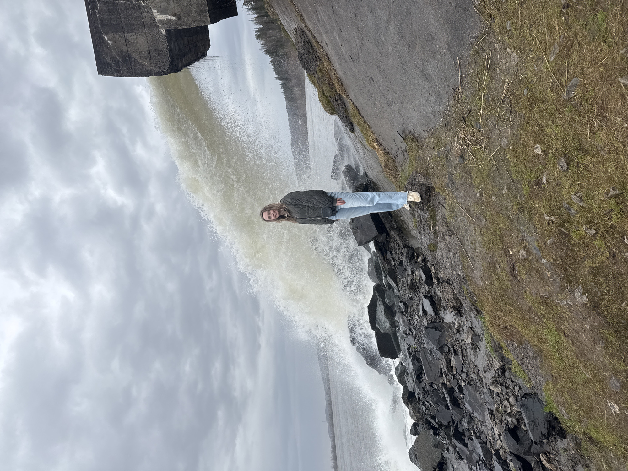 This image depicts a young woman posing next to a powerful waterfall or dam spillway in an outdoor setting, likely in Chernushka, Russia. The sky is overcast with grey clouds, suggesting a cloudy day.
A massive, dark grey concrete structure, possibly part of a dam or industrial facility, is prominently visible in the upper left portion of the image. From beneath this structure, a large volume of turbulent, light-colored water cascades downwards with considerable force, creating a wide, frothy spray.
To the right of the gushing water, a young woman is standing on a rugged bank composed of dark rocks and scattered patches of grass. She has long, light brown hair and is dressed in a dark green jacket, light blue jeans, and light-colored sneakers. The woman is smiling and looking directly towards the camera, indicating she is being photographed.
Beyond the immediate cascade, a wider body of water, likely a river or reservoir, stretches into the background. A distant treeline is faintly visible along the far shore. The ground to the right of the woman is covered with sparse grass and dry plant stalks, and a muddy path is visible near the edge of the rocks. No visible text is present in the image. The scene captures the raw power of the water against a man-made structure, with a cheerful human element.